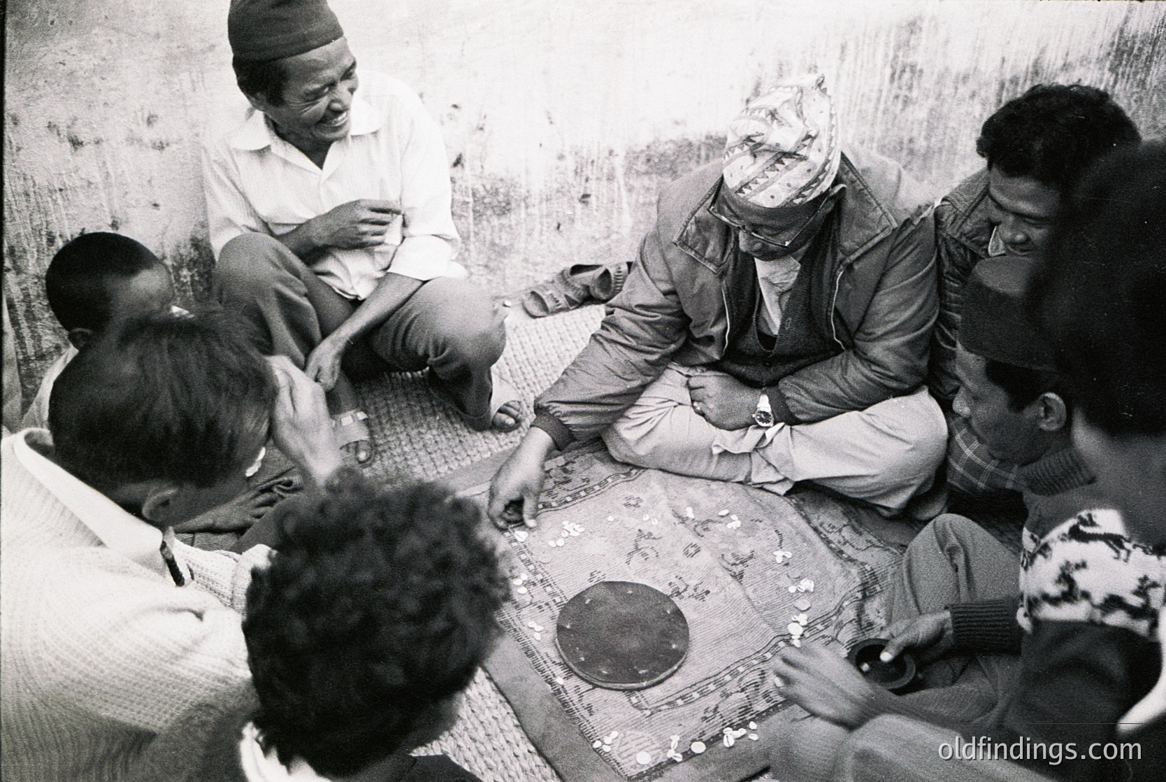 Group of men seated on woven mats in a dimly lit, textured interior, playing a traditional board game. Central figure wears a headscarf and glasses, surrounded by others in casual attire. Mid-20th century African setting, likely or . Rich cultural/historical context for anthropology studies.