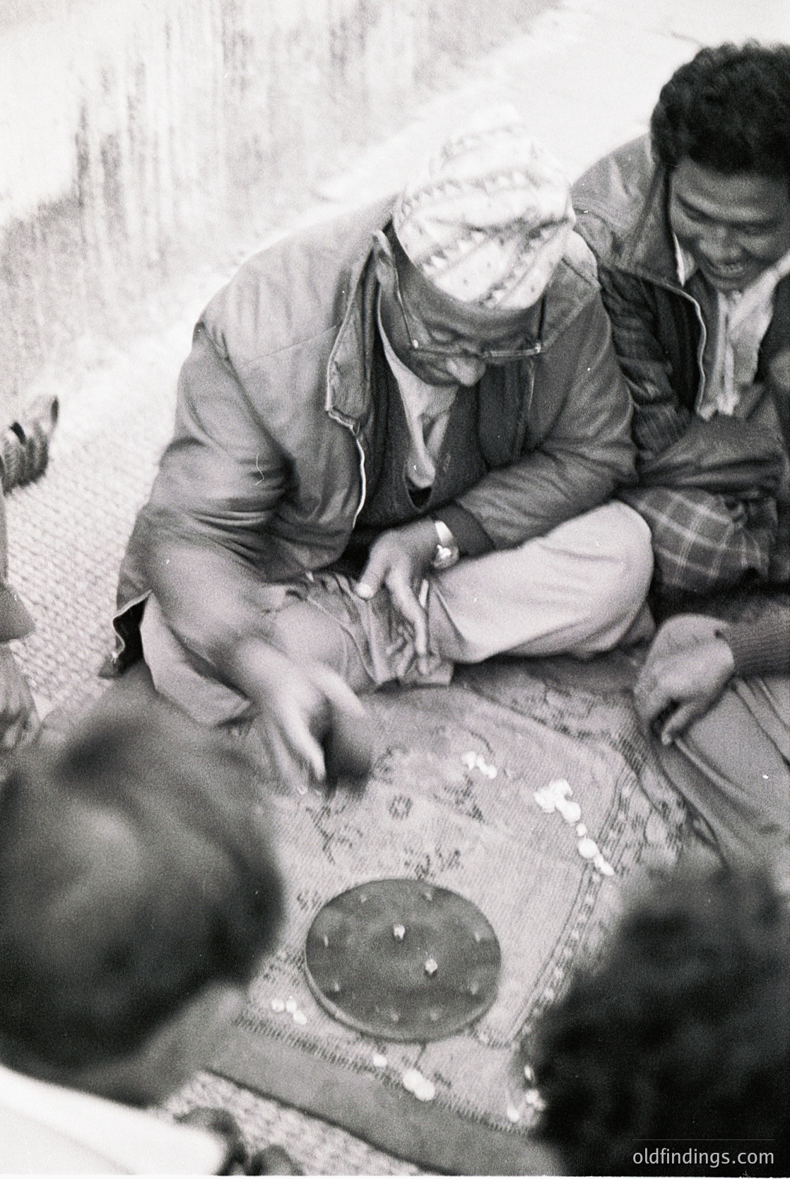 Two men in traditional headscarves and layered clothing play a board game on a woven mat, likely in a Middle Eastern or North African setting. The game board features circular markers and a central circular piece, suggesting a strategy game like backgammon. The attire and setting suggest a 1960s–1980s timeframe.