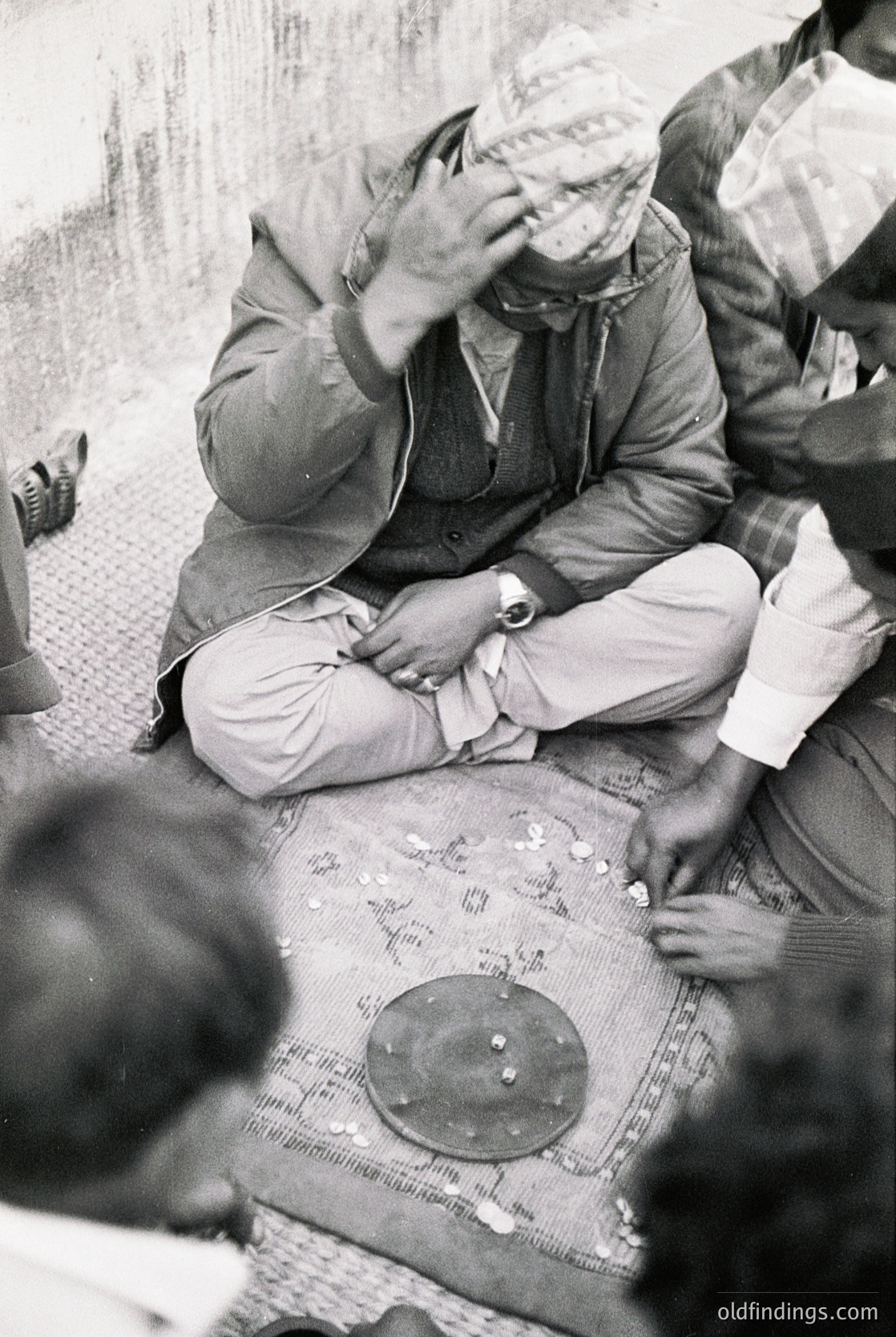 Vintage black-and-white photo of a seated man in traditional attire playing a round frame drum (*darbuka*) surrounded by others in a communal setting. Clothing suggests Middle Eastern or North African cultural influences. Likely 1950s–1970s based on style and grain.