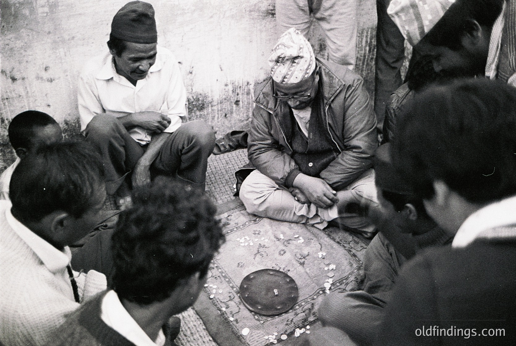 A group of men in traditional attire gathers indoors, seated on a woven mat around a central table. One man, wearing a headscarf and dark jacket, appears to be performing a ritual or ceremony with hands extended toward a circular object on the table. The setting suggests a communal or cultural practice, possibly in South Asia. The black-and-white tone and clothing styles suggest a mid-20th-century timeframe. [Traditional South Asian ritual gathering, mid-20th century ]