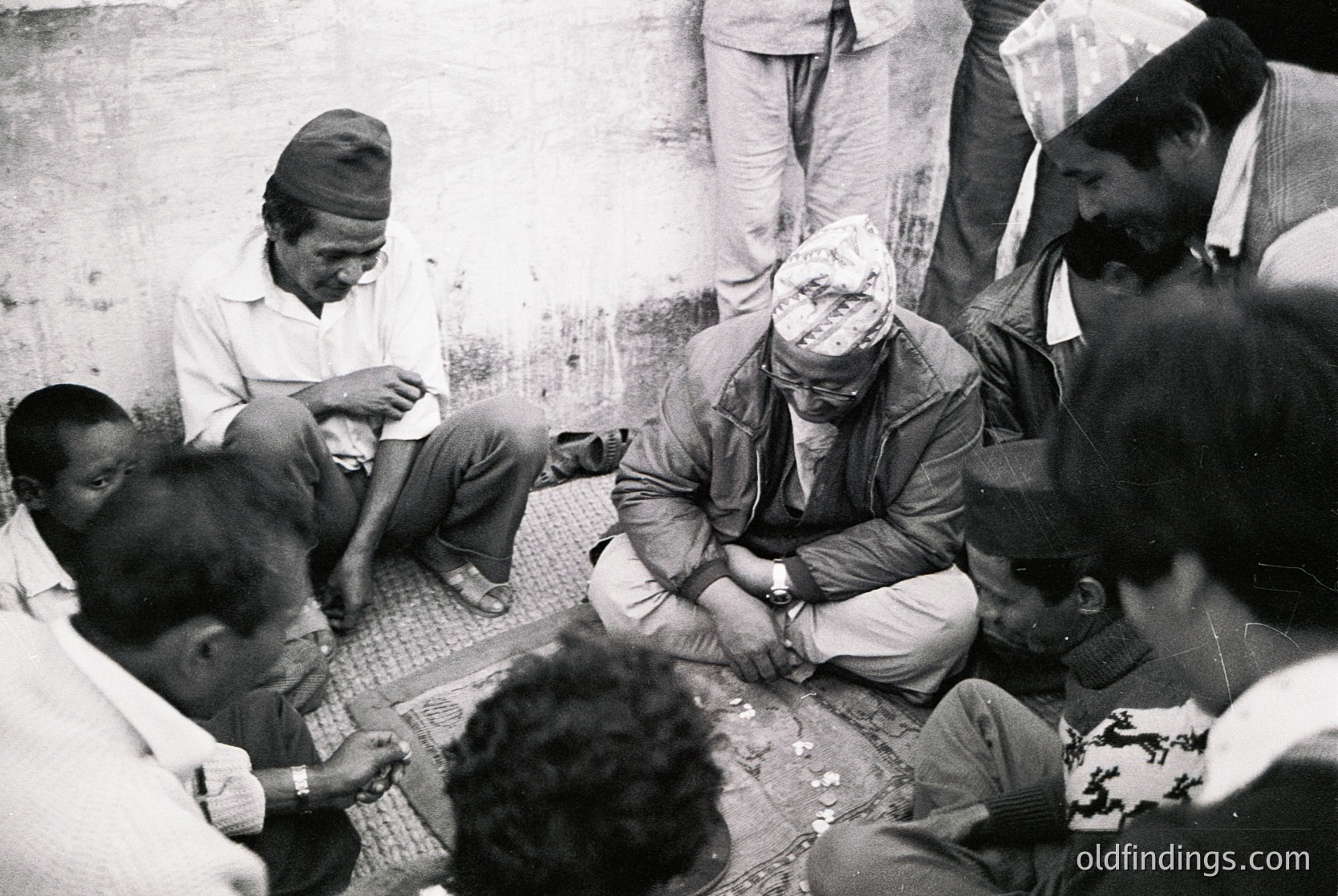 A group of men in traditional attire gathers indoors, seated on a tiled floor around a central figure wearing a headscarf and glasses. The setting appears rustic, with worn walls and a low-ceilinged space. Likely mid-20th century, possibly South Asia.