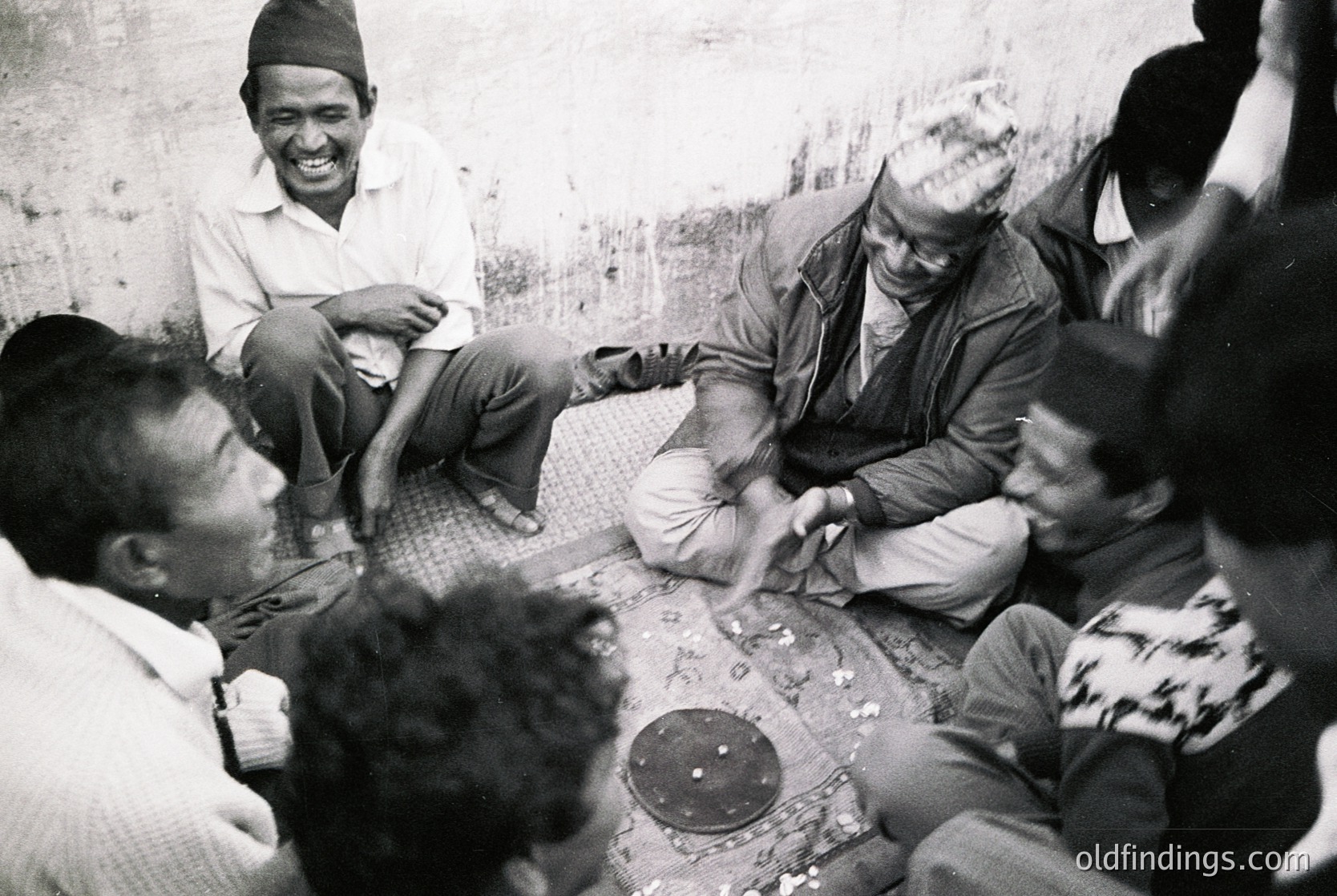 Group of men gathered in a casual, informal setting, likely Nepal, mid-20th century. Traditional attire includes headgear and loose garments; one man plays a stringed instrument. Rustic, earthen surroundings suggest rural or village life.
