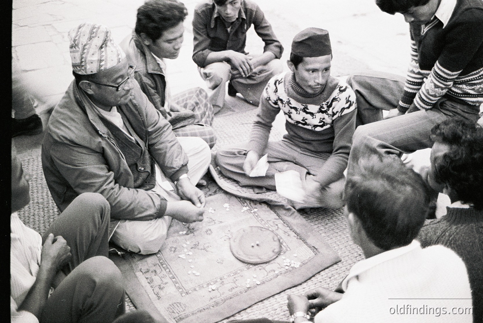 Group of men seated on floor playing traditional board game, likely in South Asia. Mid-20th century attire suggests or . Distinctive headwear and patterned textiles indicate cultural heritage. Game board features circular markers and intricate designs.