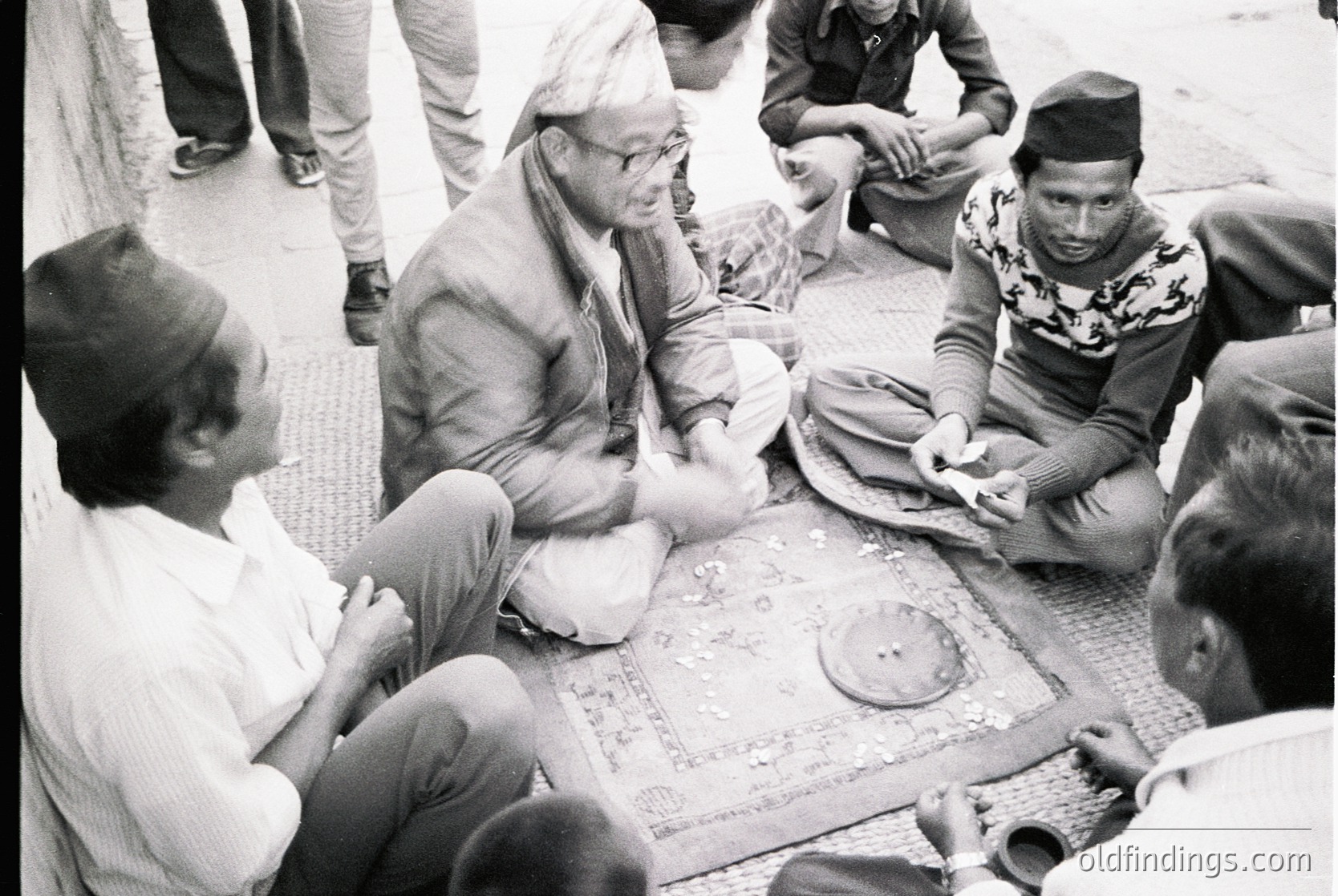 Group of men seated on patterned rugs in traditional attire, engaged in a communal activity—likely a card game or discussion—outdoors. Clothing includes headgear (e.g., *pagri*) and long-sleeved garments, suggesting South Asian cultural context. Mid-20th century (1950s–1970s) based on style and black-and-white format.