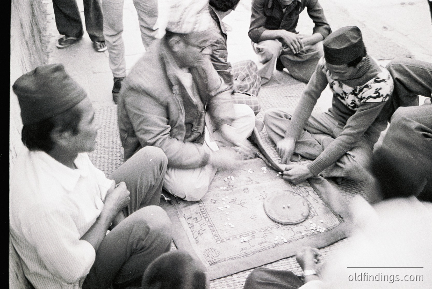 Group of men playing traditional board game on patterned rug, mid-20th century. Indoor setting with blurred background, suggesting communal focus. Clothing includes headwear and casual attire.