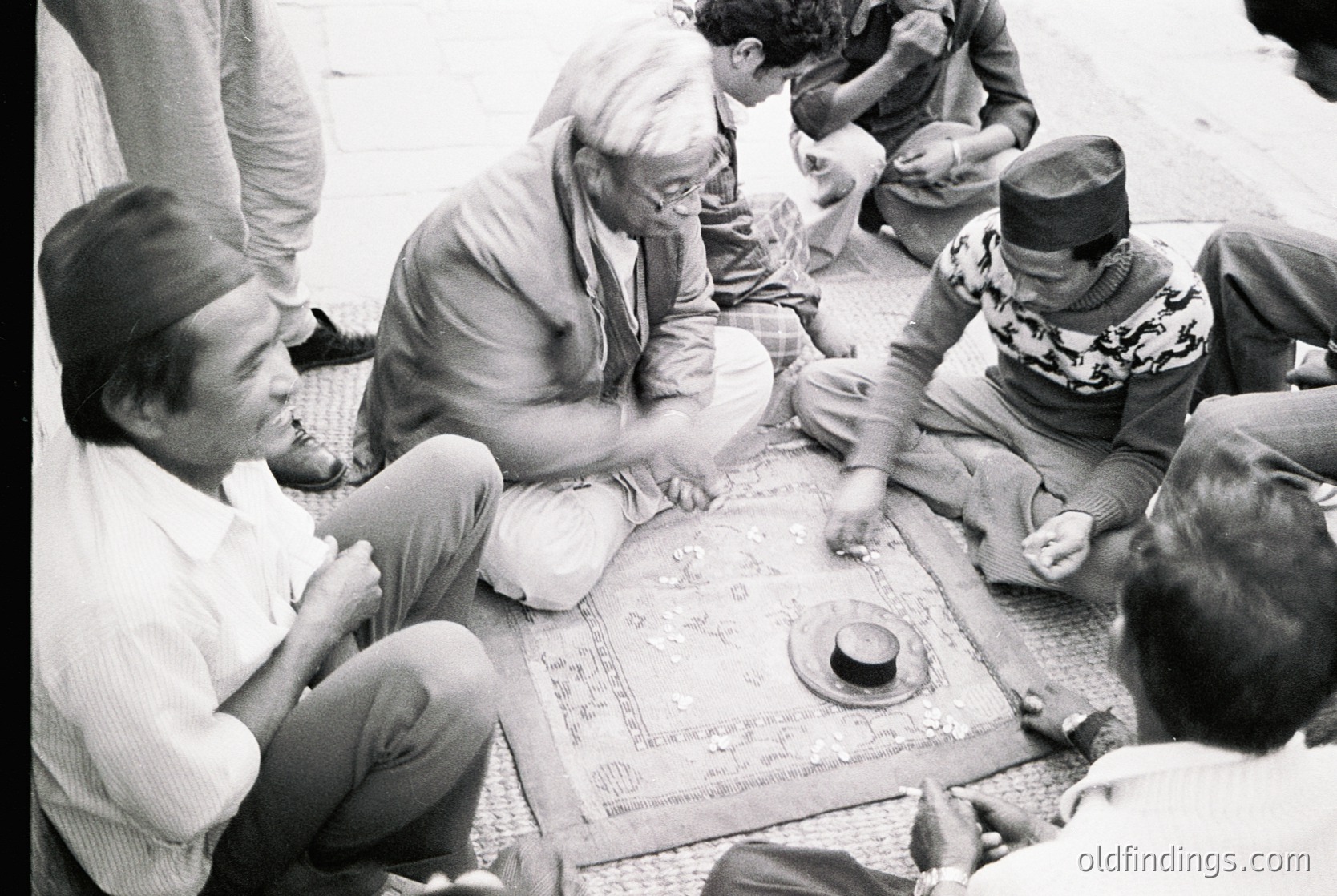 Group playing traditional board game on woven mat, likely backgammon, in 1960s-70s Middle East. Men in traditional headwear and casual attire gather around a wooden board with Arabic script. Informal, communal setting captures cultural heritage and social bonding.