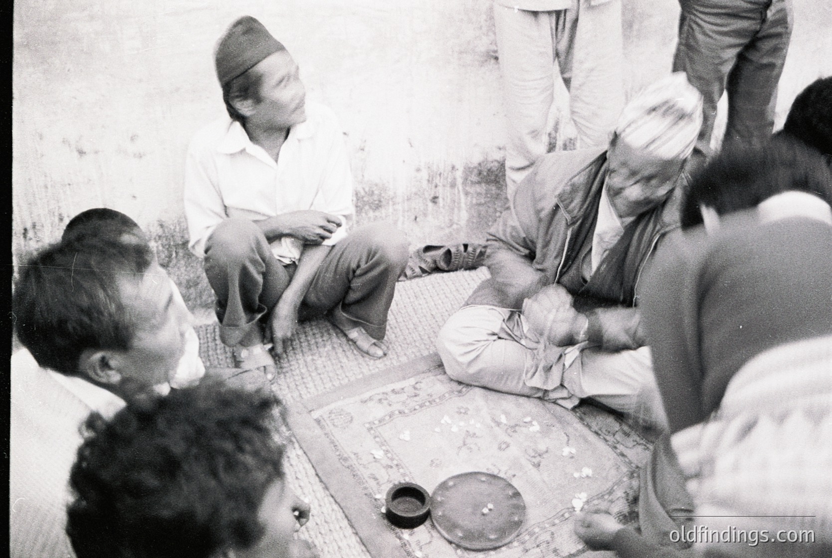 Group of men seated on patterned rugs in a traditional indoor setting, likely a home or communal space. One man wears a turban, another holds a small metal bowl. Mid-20th century attire and setting suggest cultural or familial gathering. Possible South Asian region.