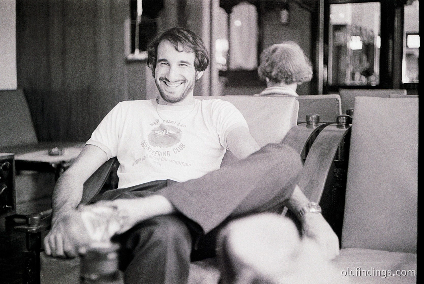 Mid-century barber shop interior with a relaxed man in a vintage "Every Living Thing" T-shirt, seated in a classic upholstered chair. Barber pole and vintage styling tools visible in background. Likely 1970s USA.