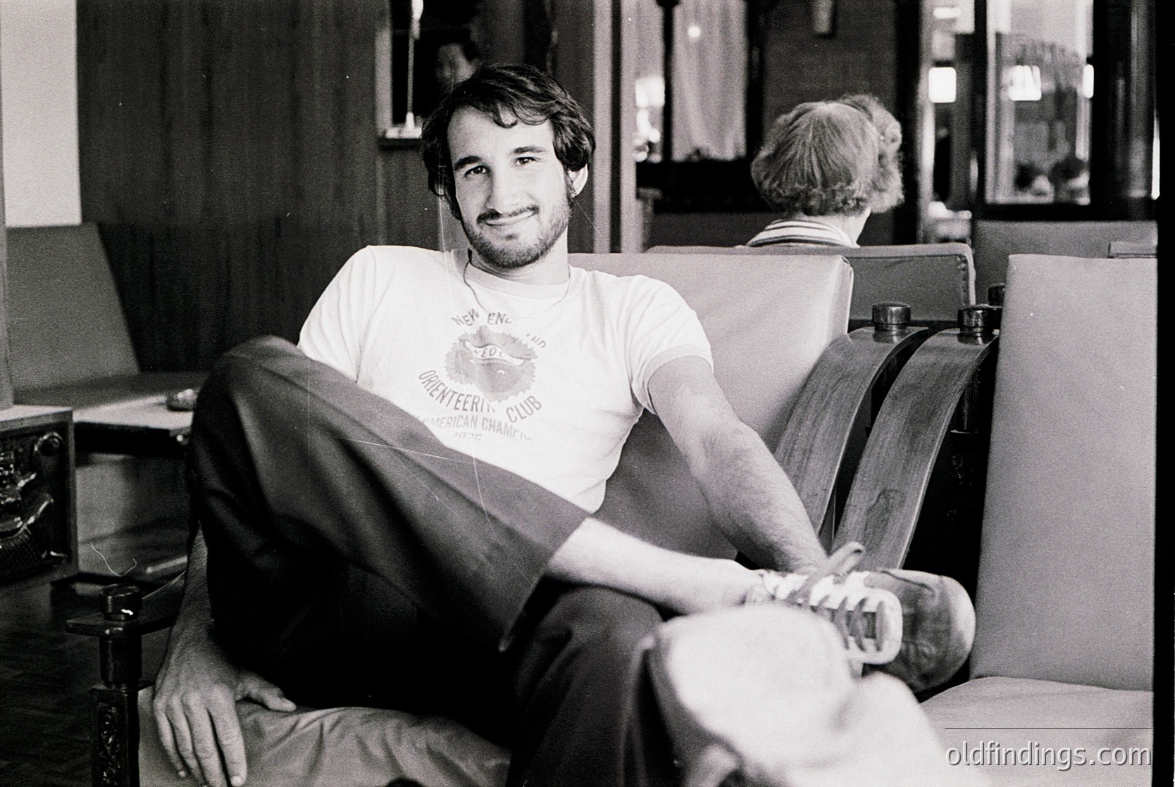 Black-and-white portrait of a relaxed man in a 1970s-style T-shirt ("Keep Peace & Quiet" with a peace symbol) lounging on a leather couch in an indoor setting. Mid-century furnishings and a typewriter in background suggest an office or creative workspace.