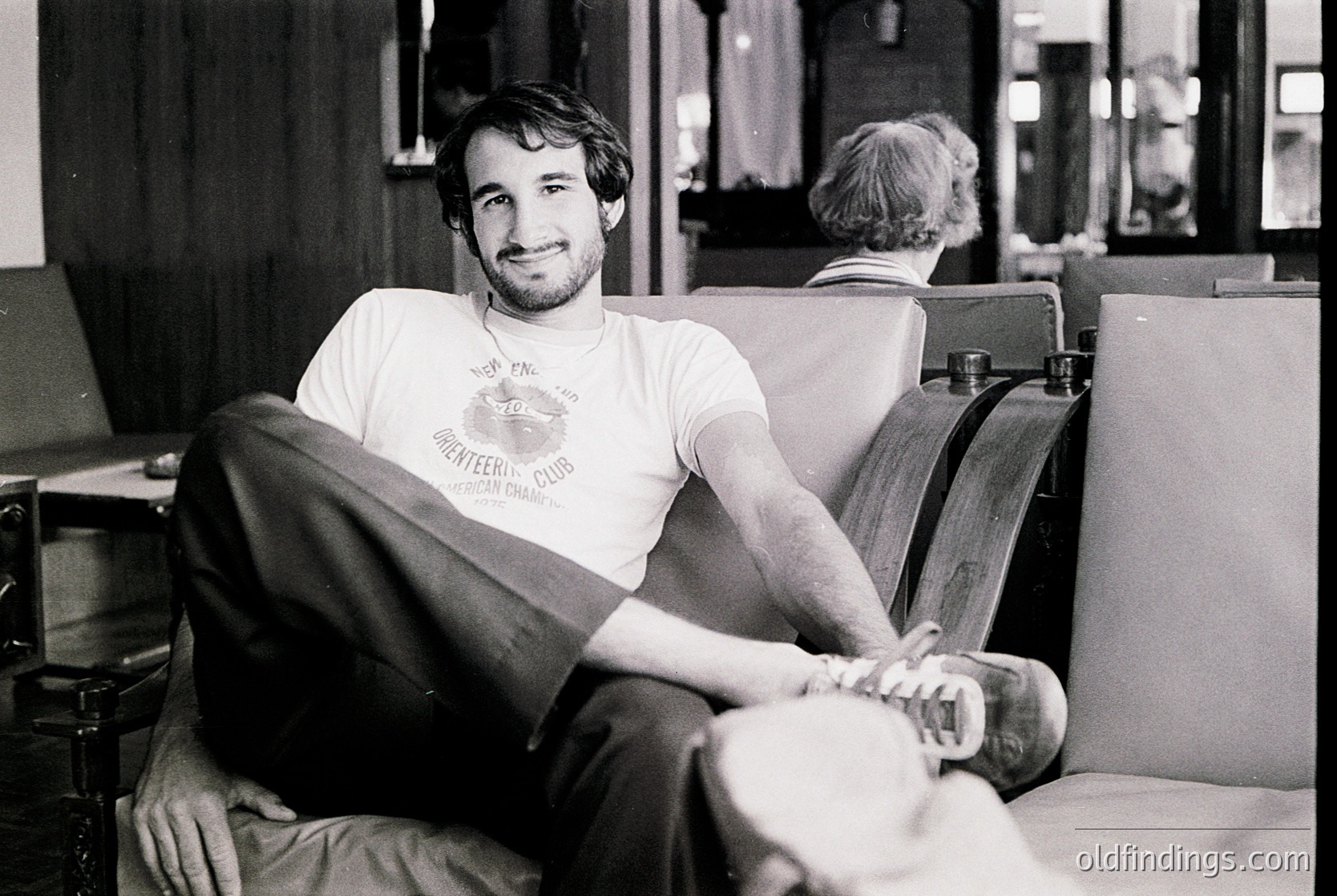 Relaxed man in a 1970s-style T-shirt (with "Greenwich Village" and "Scout Club" text) lounges on a leather armchair in an indoor setting, possibly a café or lounge. Blurred background suggests a social, mid-century environment. Black-and-white composition highlights candid, informal vibes.