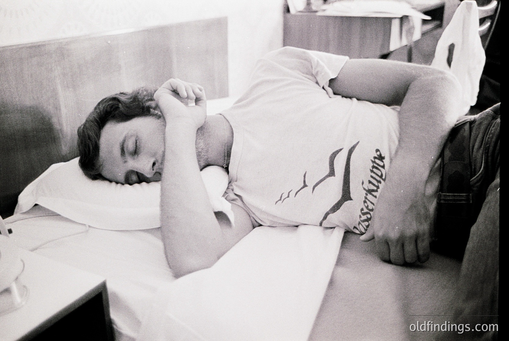 Black-and-white candid of a man resting on a bunk bed, shirtless with a "Lacoste" logo on his shorts. Minimalist dorm-style interior with a desk and chair in background. Likely 1970s–1980s, Western Europe/US. Evokes nostalgia for student life or travel.