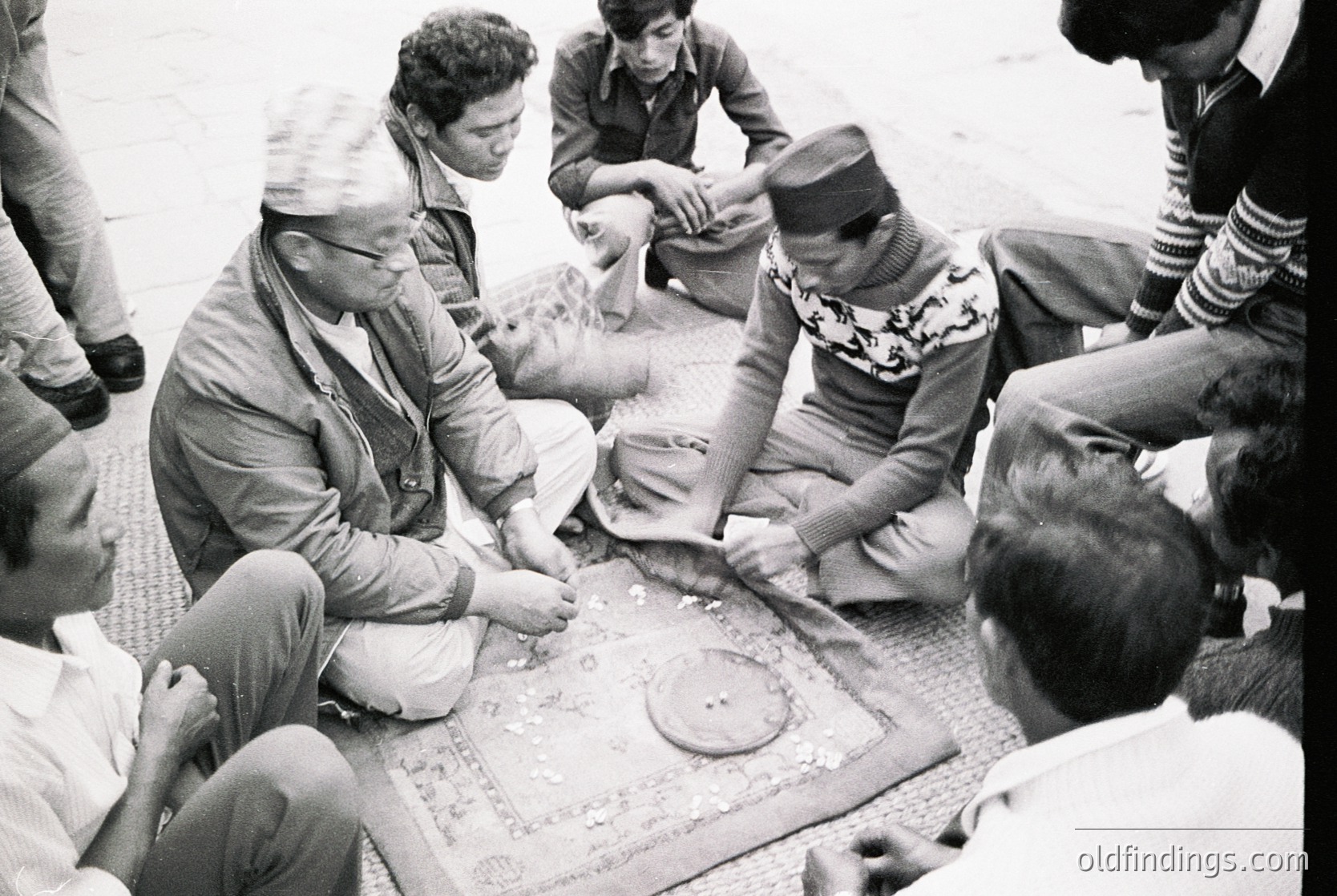 Black-and-white photo of a group playing a traditional board game in a communal setting, likely . Men in traditional attire (headscarves, long robes) and casual wear gather around a large, ornate game board on the ground. One man holds a small animal, possibly a goat, adding cultural context. Urban or courtyard environment with visible architectural elements like columns.