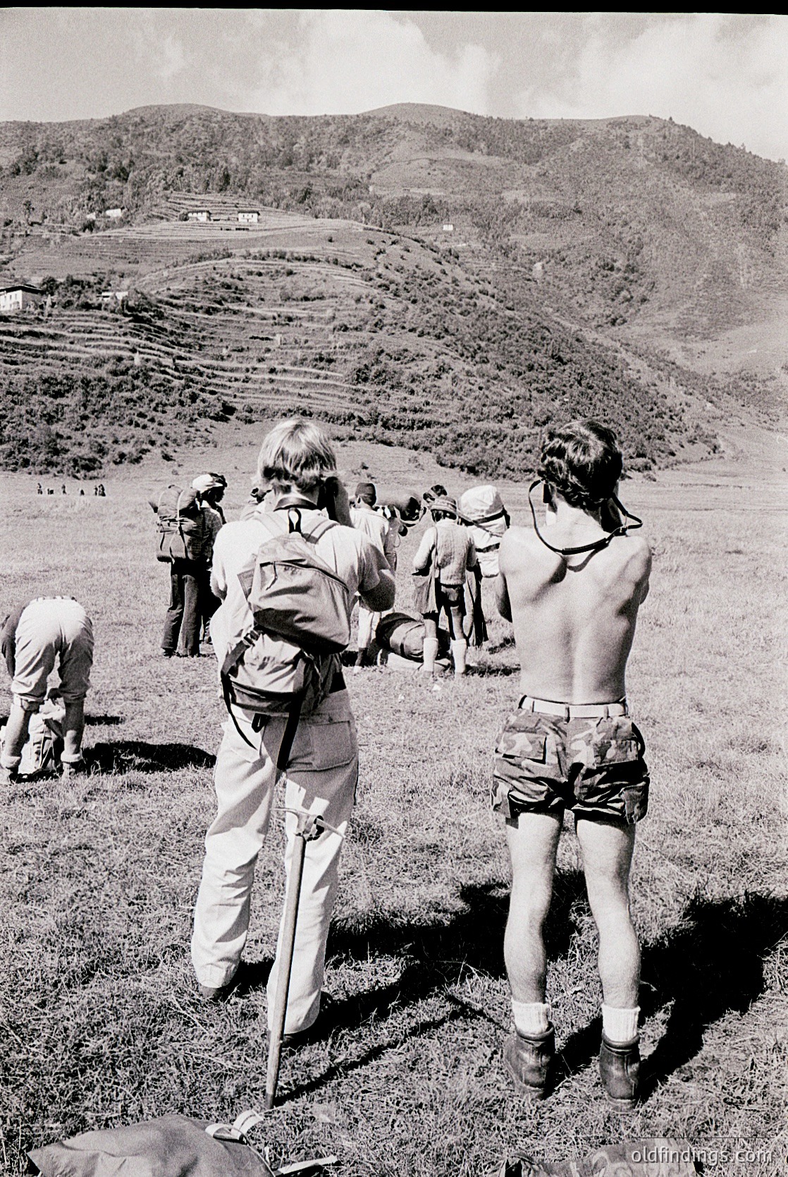 Group of hikers in 1970s-style outdoor gear stands on a terraced hillside, likely in a Mediterranean or alpine region. Shorts, backpacks, and hiking poles suggest a guided trek. Mid-century hiking culture and rugged landscapes depicted.
