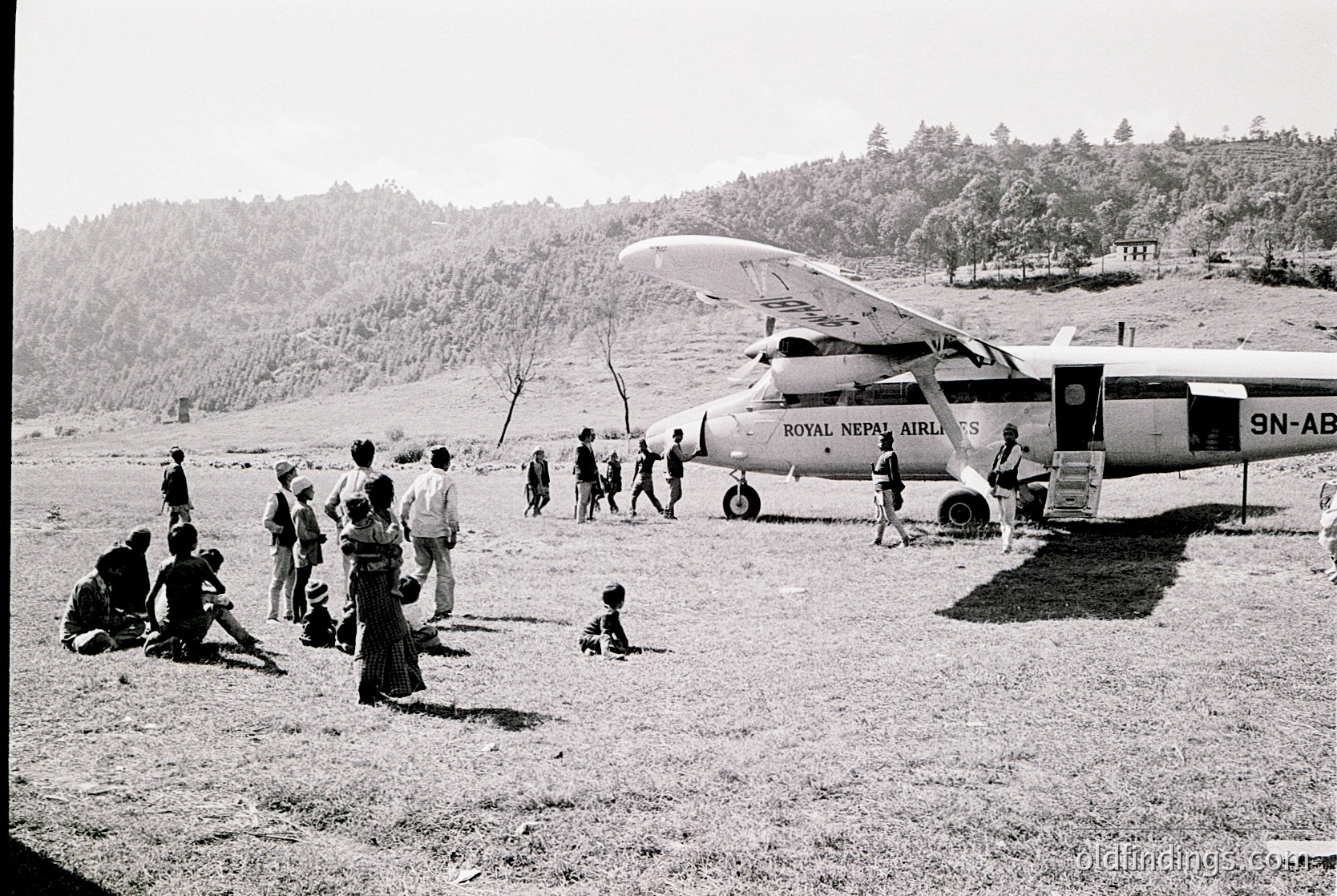 A group of people gathers around a Royal Nepal Airlines floatplane (registration 9N-ABX) on a grassy airstrip, likely in the Himalayan region. The aircraft’s seaplane design suggests remote, high-altitude operations. Mid-20th century attire and the plane’s livery indicate the 1960s–1970s era.