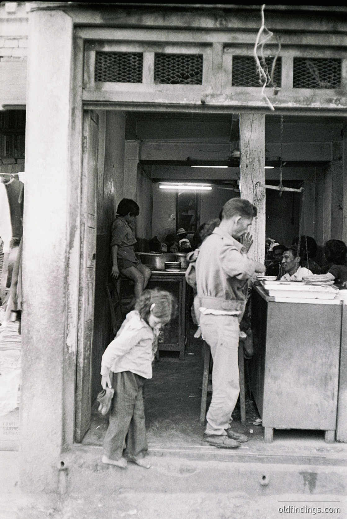 Vintage street-side café or snack bar interior, likely mid-20th century. A man in a light apron serves food from a counter, while a child in a white blouse and shorts stands nearby. Wooden beams and exposed wiring add to the rustic, utilitarian atmosphere. Crowded, informal seating with tables and chairs visible inside. é