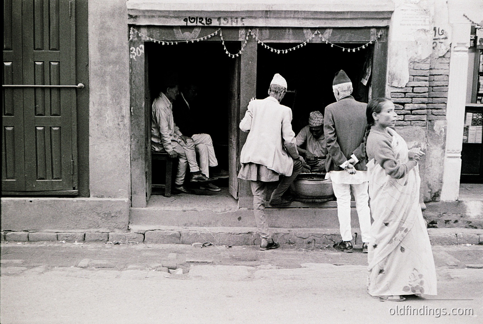 Black-and-white street scene in Nepal, likely Kathmandu, mid-20th century. Traditional attire—men in *dhaka* and *topi*, women in *sari*—gathers outside a wooden doorway adorned with festive garlands. A seated man in the doorway holds a pipe, while a woman in white sari carries a tray. Brick-paved street and stone steps enhance cultural authenticity.