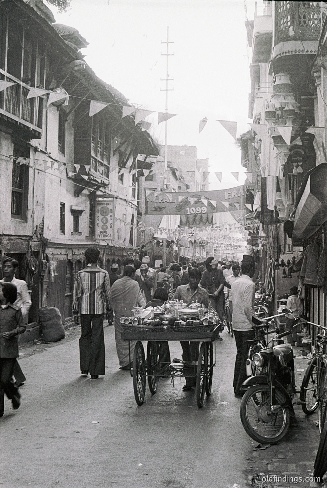 Vibrant street scene featuring a bustling market cart laden with fresh produce, likely melons and vegetables, in a narrow alleyway. Decorative flags and signs adorn buildings with colonial-era architecture, hinting at a historic urban setting. Crowds of casually dressed pedestrians and a motorbike suggest mid-20th-century daily life, possibly in a Mediterranean or Middle Eastern locale.