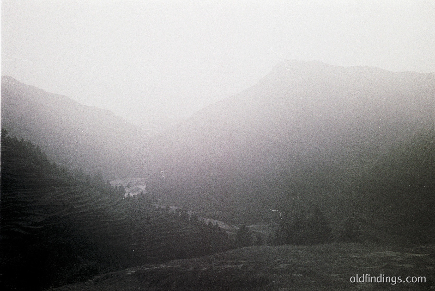 Misty mountain valley with terraced fields and sparse structures, captured in monochrome. Fog obscures distant ridges, emphasizing depth. Likely rural agricultural landscape, possibly East/Southeast Asia.