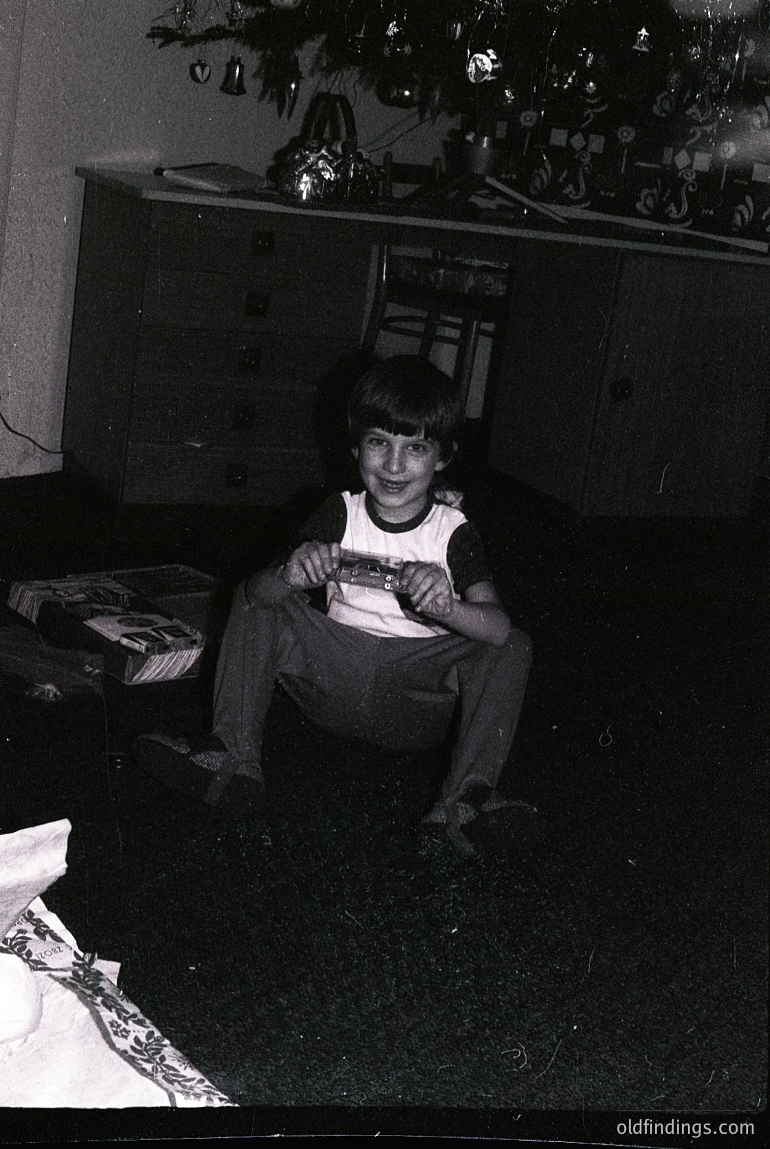 Vintage black-and-white photo of a young boy in a 1970s-style tank top and pants, holding a wrapped gift under a decorated Christmas tree. Wooden cabinet and wrapped presents in background suggest holiday cheer. Warm, nostalgic family moment.