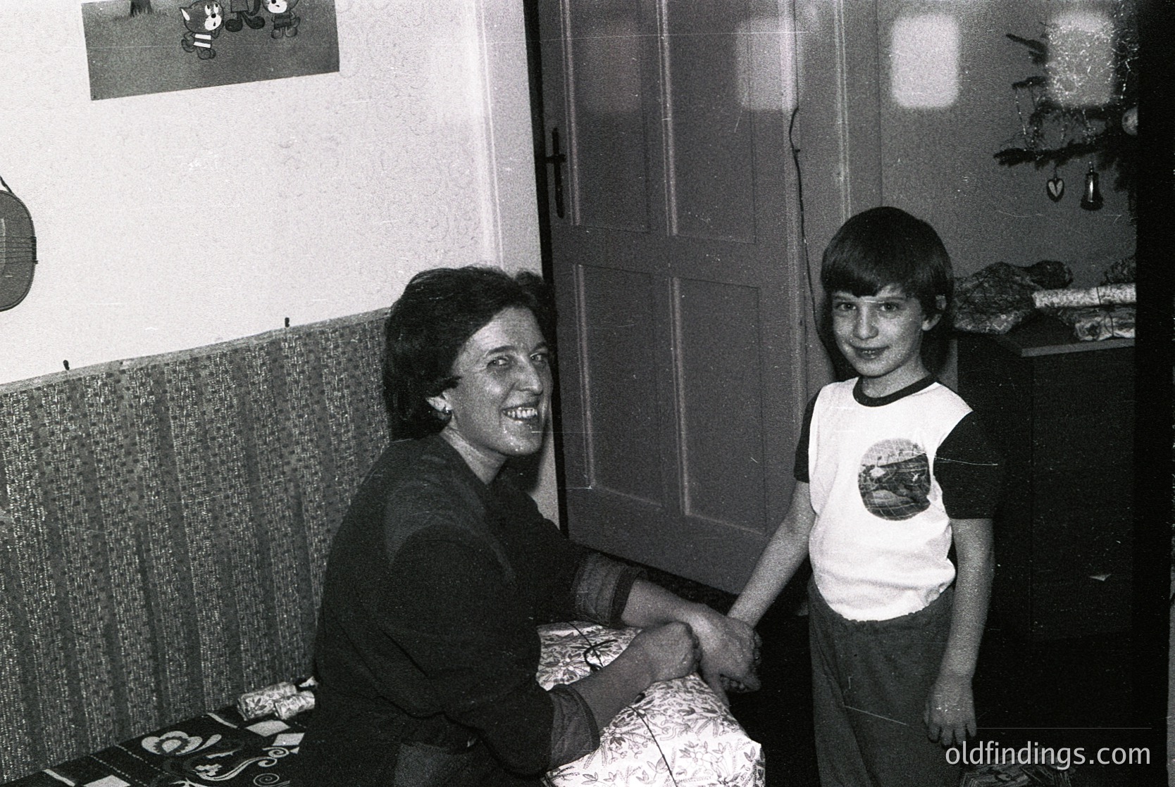 Black-and-white indoor portrait of a woman and young boy shaking hands, likely mid-1970s–1980s. Woman wears a dark blouse with a pearl necklace; boy in a white T-shirt with a graphic print and rolled pants. Background shows a curtain, framed art, a Christmas tree, and a wooden door. Warm, candid family moment.