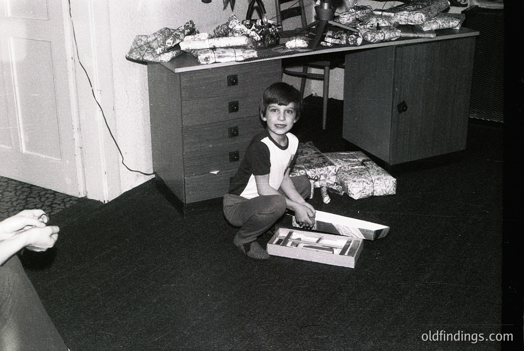 Mid-century modern wooden dresser with stacked towels atop, 1960s-70s interior. Boy kneels on dark carpet, opening a cardboard box with scissors. Retro household setup reflects mid-century design.