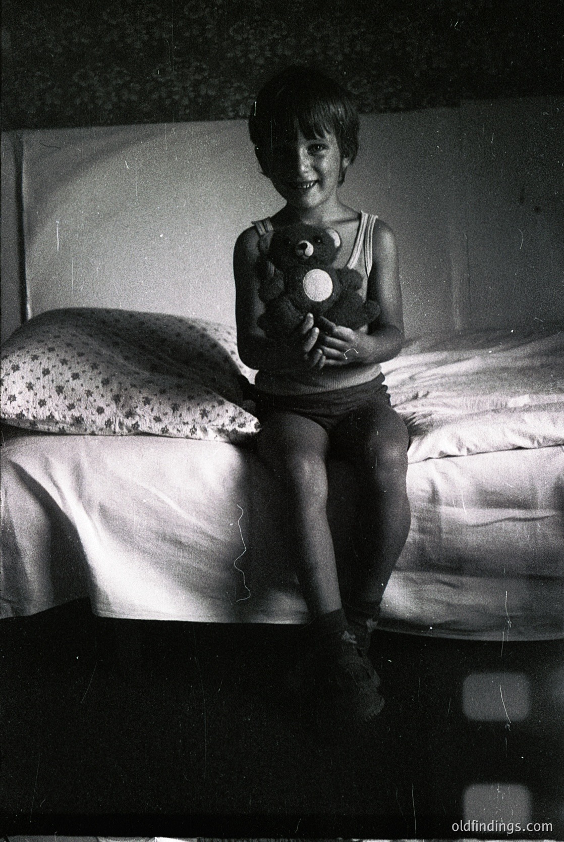 A young child sits on a twin bed holding a stuffed toy, likely from the 1950s–1960s. The room features floral-patterned bedding and a simple wooden headboard. The vintage black-and-white tone suggests mid-century domestic life.