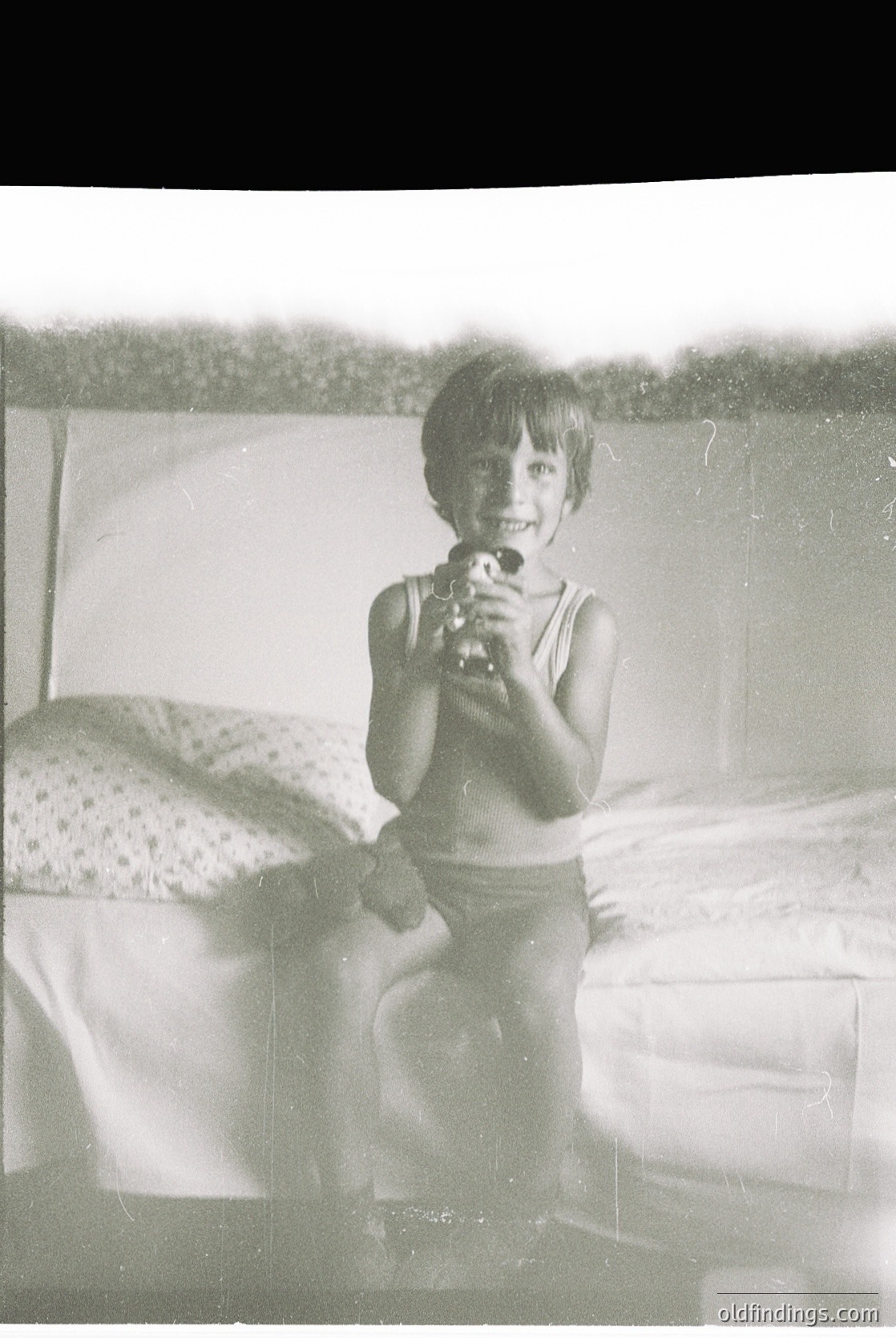 Vintage black-and-white photo of a child holding a toy gun, likely a 1950s–1960s indoor portrait. Patterned bedding and partial curtain suggest mid-century domestic setting. Candid, nostalgic snapshot captures youthful innocence.