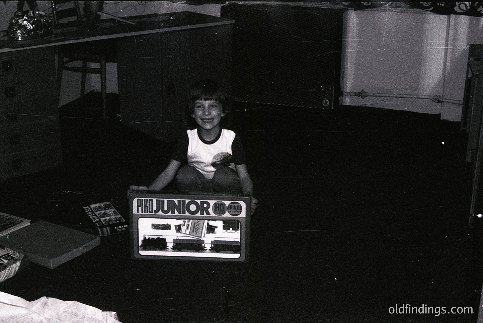 Child poses with a vintage **Junior OO gauge model train set** (1960s-70s) in a dimly lit indoor setting. Noticeable details: wooden shelves, cardboard boxes, and a curtained window. Evokes mid-century toy nostalgia.