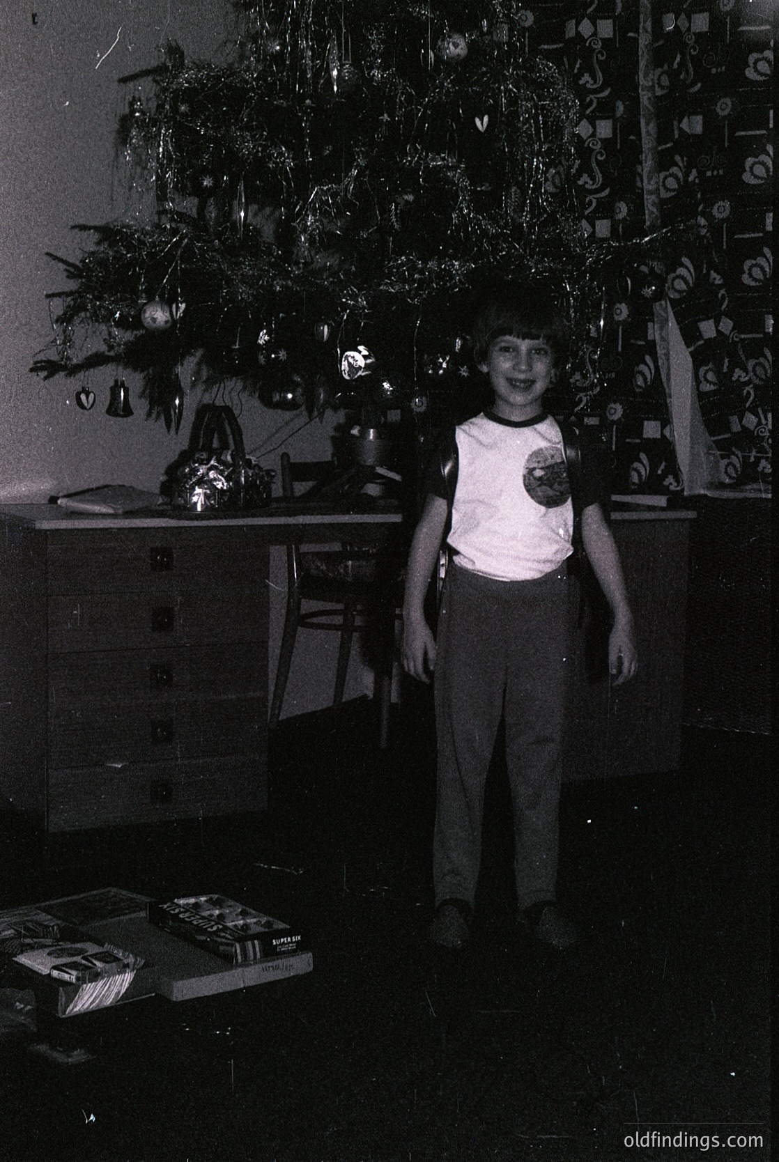 Vintage indoor holiday scene featuring a young child in 1970s-style clothing (short-sleeve shirt, pleated pants) standing beside a decorated Christmas tree adorned with tinsel, baubles, and paper ornaments. Wooden dresser with stacked books and framed artwork in background. Warm, dim lighting enhances nostalgic atmosphere.