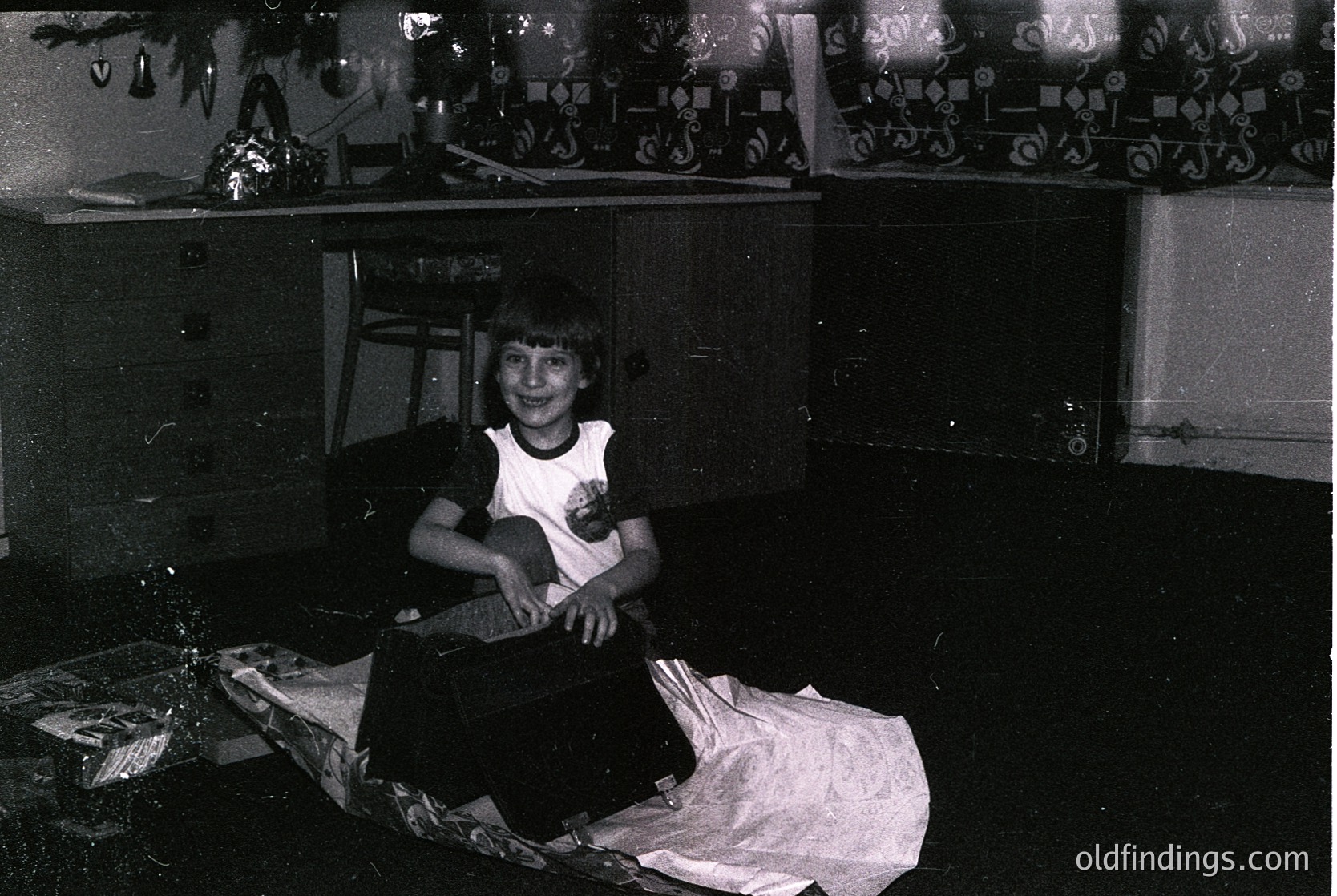Black-and-white interior shot of a person seated on a bed in a modest, cluttered room. Decorative wallpaper with geometric patterns frames the scene. The individual wears a white tank top with a small graphic and dark pants. Shelves above display mismatched items, including a framed poster. Likely mid-20th century domestic setting.