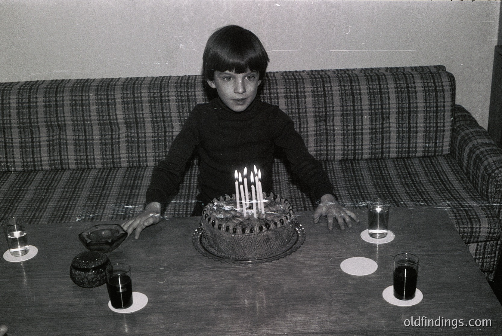 Young boy in 1970s-style sweater poses with birthday cake adorned with 11 lit candles on a dark wooden table. Plain checkered sofa and simple glassware suggest mid-century home decor. Candid moment captures innocence and celebration.