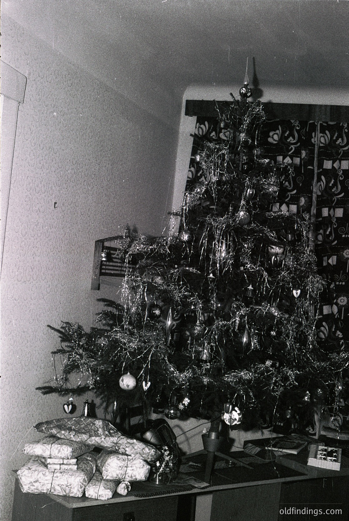 Mid-century Christmas tree adorned with tinsel, paper ornaments, and a star topper, placed beside wrapped gifts and a shelf of decorative items. Likely 1950s–1960s domestic interior, evoking retro holiday nostalgia.
