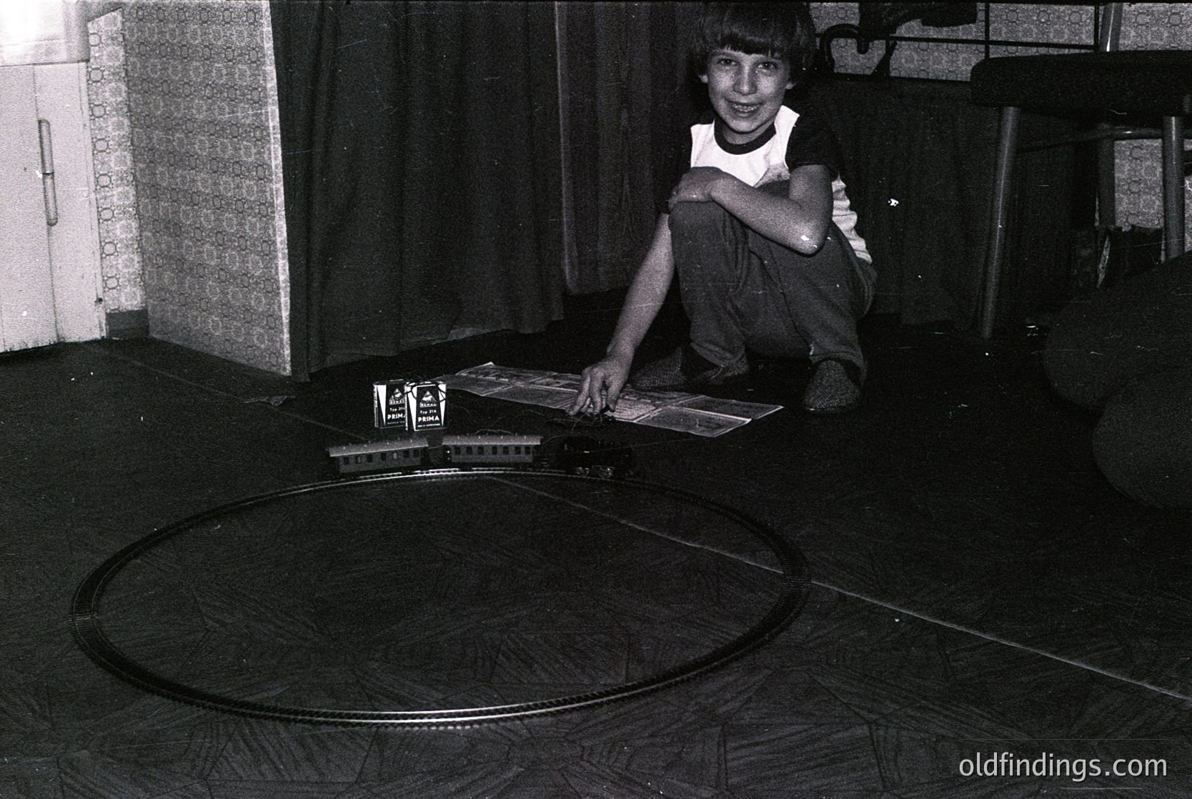 Mid-20th century boy poses with vintage setup on a circular track. Indoor setting features patterned wallpaper and a wooden floor. Casual attire includes a white shirt and shorts. Evokes nostalgia for mid-century childhood play.