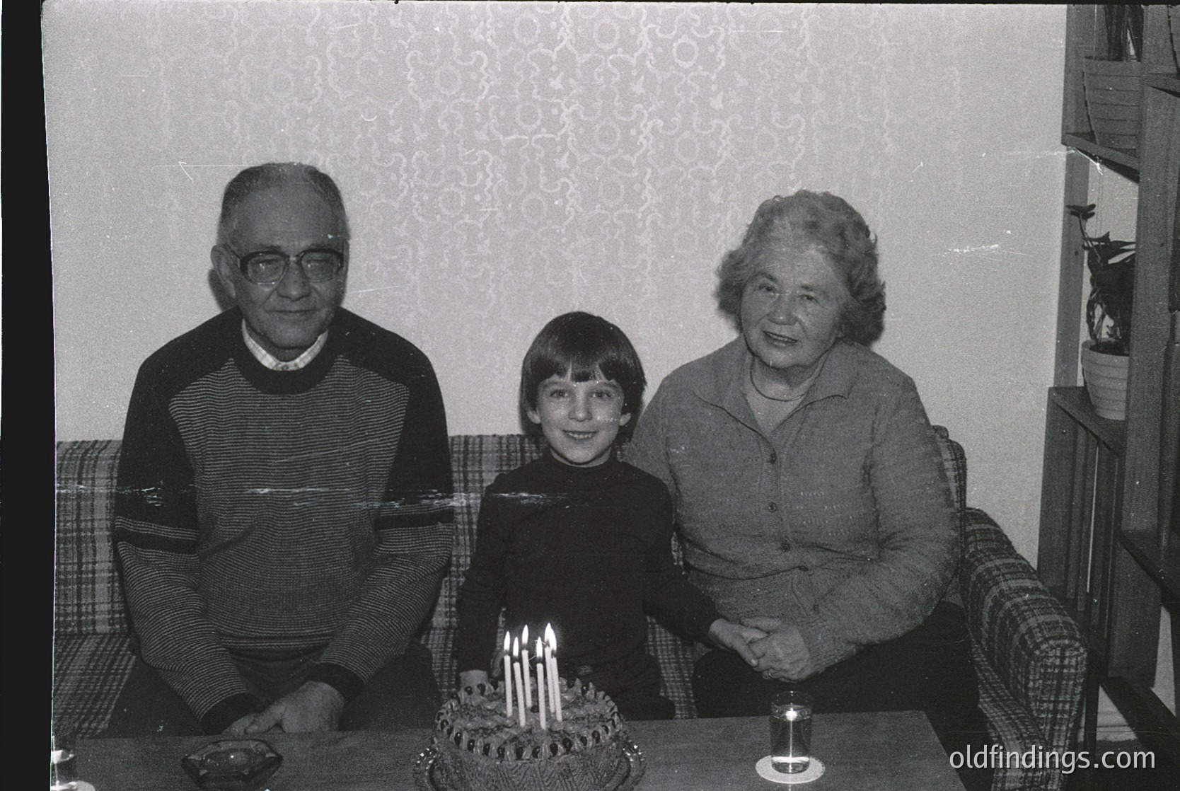 Black-and-white family portrait from the 1970s–1980s, featuring a young boy seated between two elderly adults on a patterned sofa. The boy holds a birthday cake with lit candles on a small table. Decor includes a floral wallpaper and a vintage armchair.