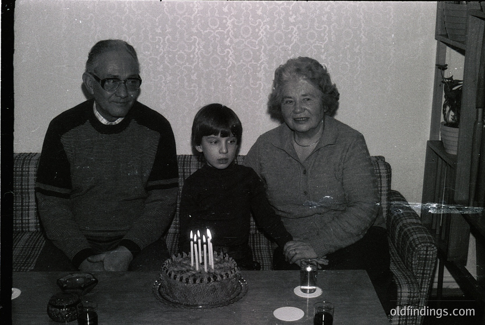 Family portrait featuring a grandfather, grandmother, and child seated at a table with a birthday cake adorned with lit candles. Indoor setting with patterned wallpaper and vintage furniture. Likely late 20th century (1970s–1980s).