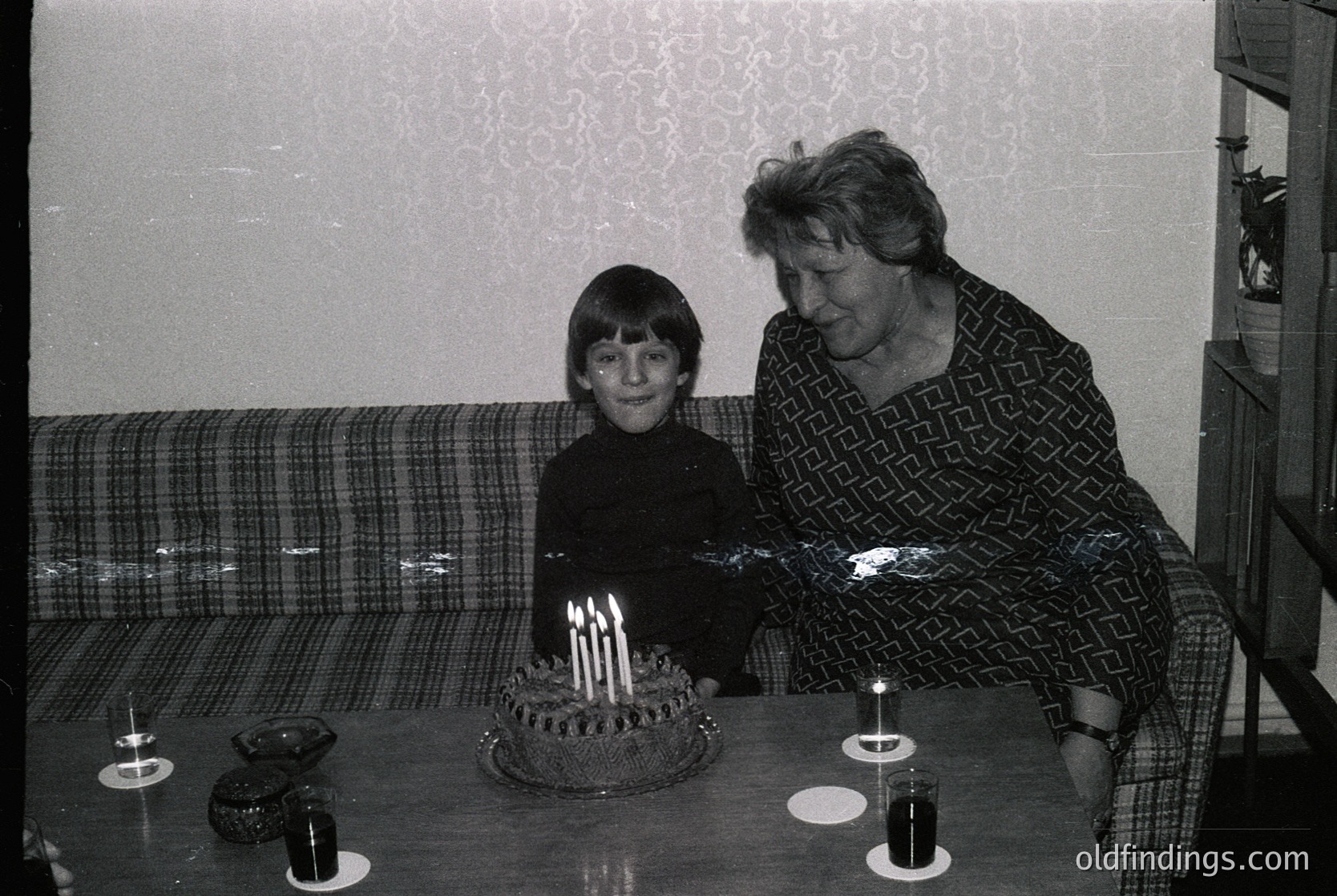 A tender 1970s family moment: a young boy and elderly woman pose beside a round birthday cake with lit candles. The woman wears a patterned blouse and skirt; the boy sits in a plaid armchair. Plain interior with minimal decor.