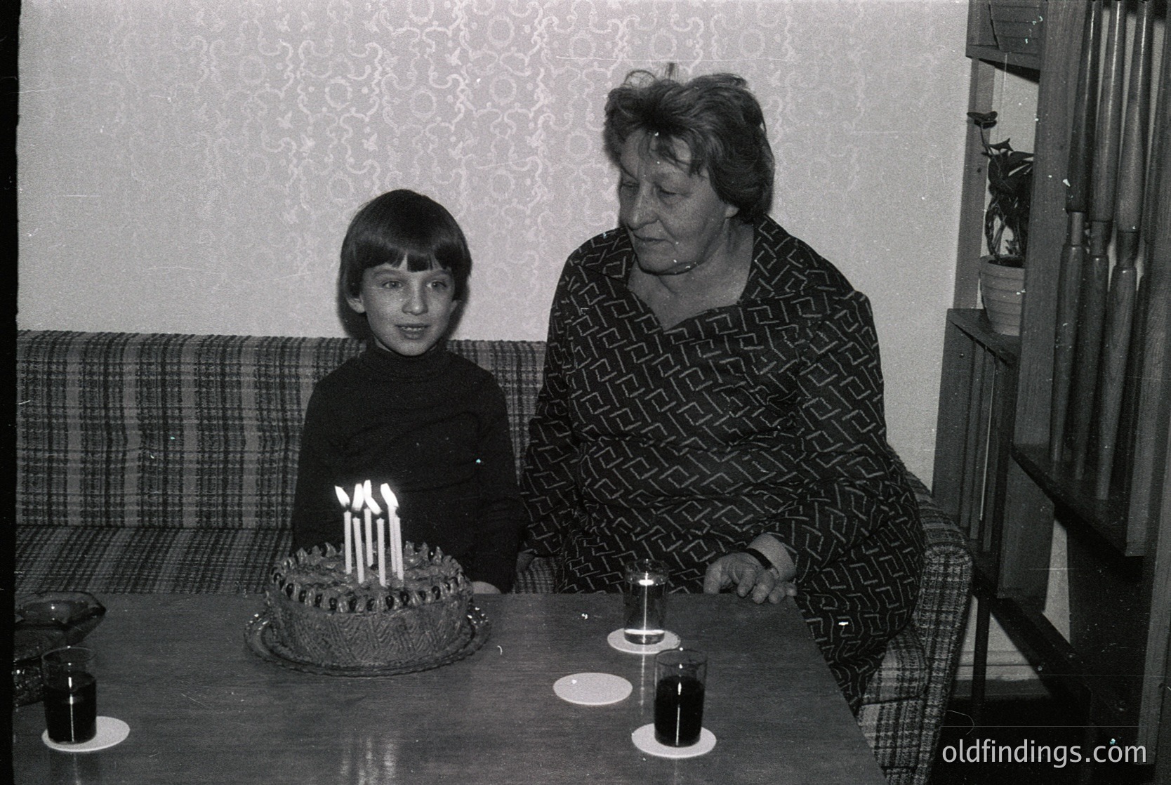 A black-and-white indoor portrait from the 1970s–1980s, featuring a grandmother and young boy at a birthday table. The boy, seated, faces a round cake with lit candles, while the grandmother stands beside him. Patterned wallpaper and a plaid sofa frame the scene, suggesting a mid-century home setting.