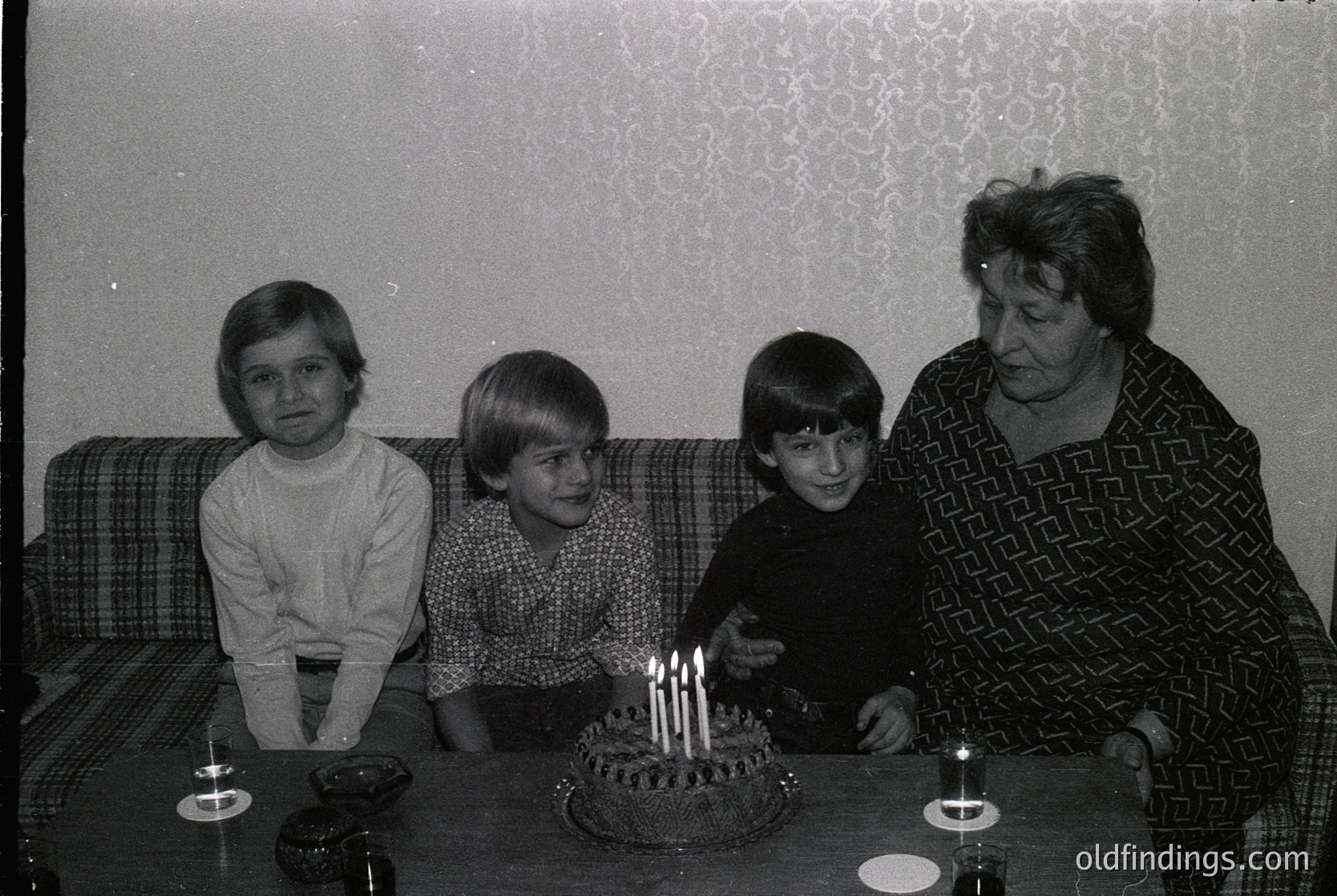 Family portrait from the 1970s–1980s, likely Eastern Europe. Three children and an adult woman pose indoors beside a round birthday cake with lit candles. The woman wears a patterned blouse, children in casual 70s-style clothing. Plain wallpaper and patterned upholstery suggest mid-century home decor.