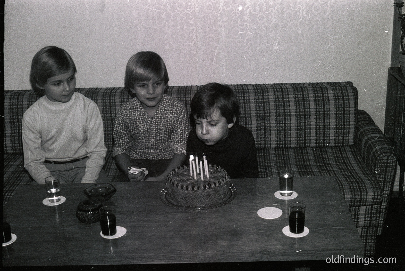Three children in 1970s-style clothing gather around a birthday cake with lit candles on a round table. Plaid sofa and patterned wallpaper suggest mid-century home decor. Small candlesticks and empty glasses flank the cake.