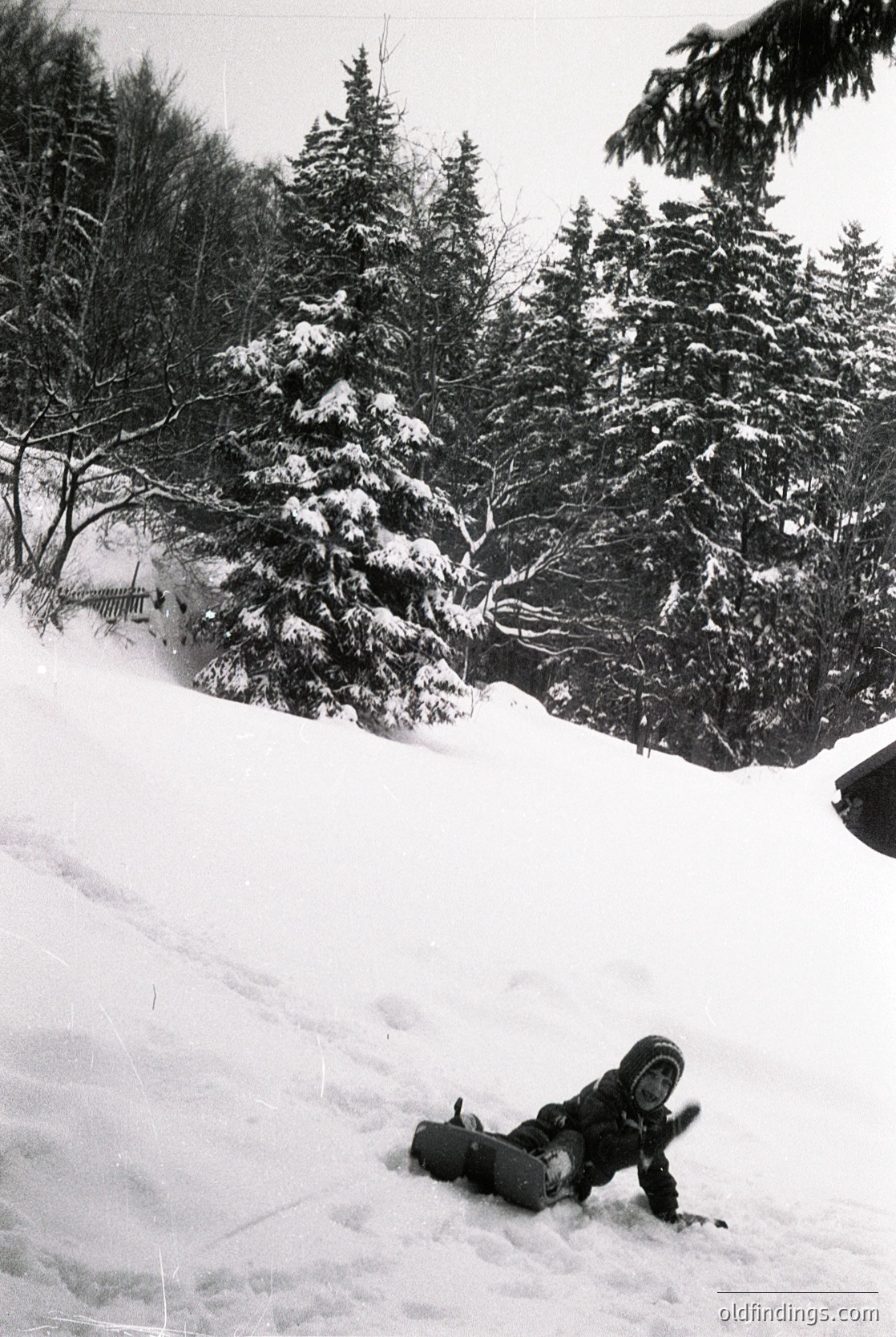Snowboarder mid-fall on groomed slope, surrounded by dense coniferous forest. Classic 1990s winter sports attire—helmet, goggles, padded jacket, and snowboard. Likely European alpine resort due to terrain and vegetation.