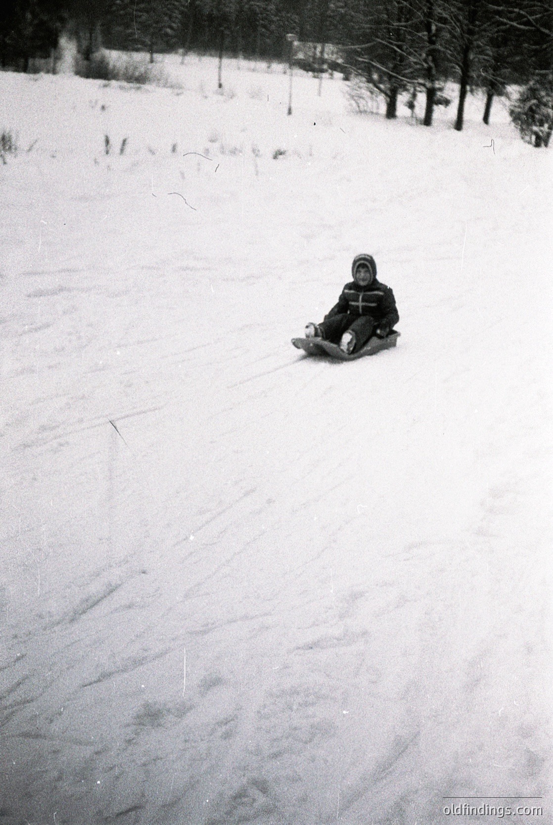 Child sledding on snow-covered slope in forested area, mid-20th century winter attire. Snow tracks indicate recent activity.