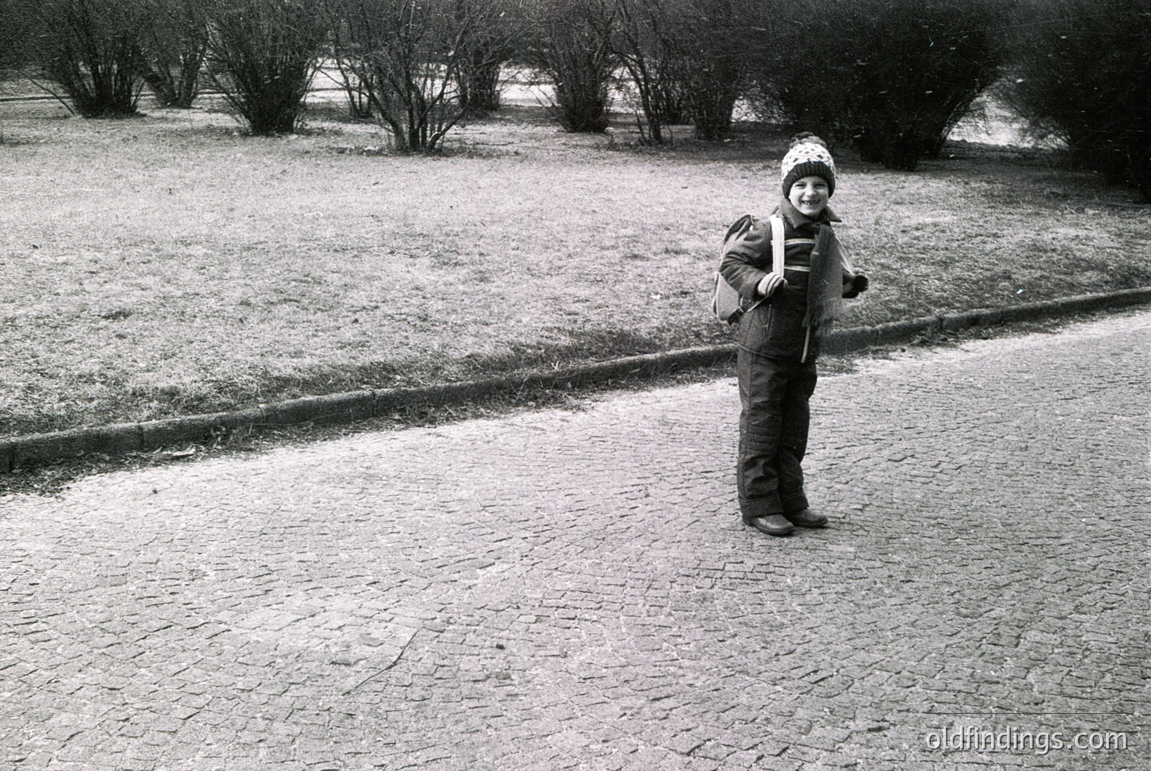Mid-20th century black-and-white photo of a child in winter attire—beanie, jacket, and backpack—standing on a cracked asphalt road. Snow covers grassy areas and tree branches, suggesting early spring or late winter. Urban park or residential setting.