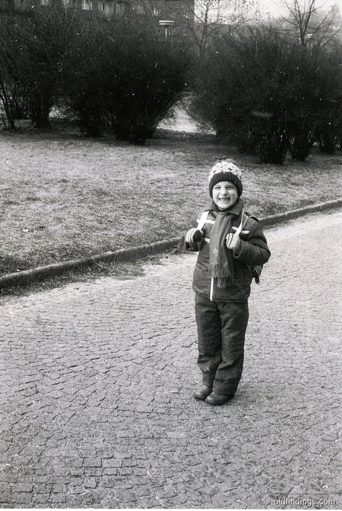 Young child in winter attire—knit hat, padded coat, and boots—stands on a cobblestone path in a park-like setting. Snowy trees and residential buildings in background suggest a cold, urban winter scene. Likely late 20th century (1970s–1990s).
