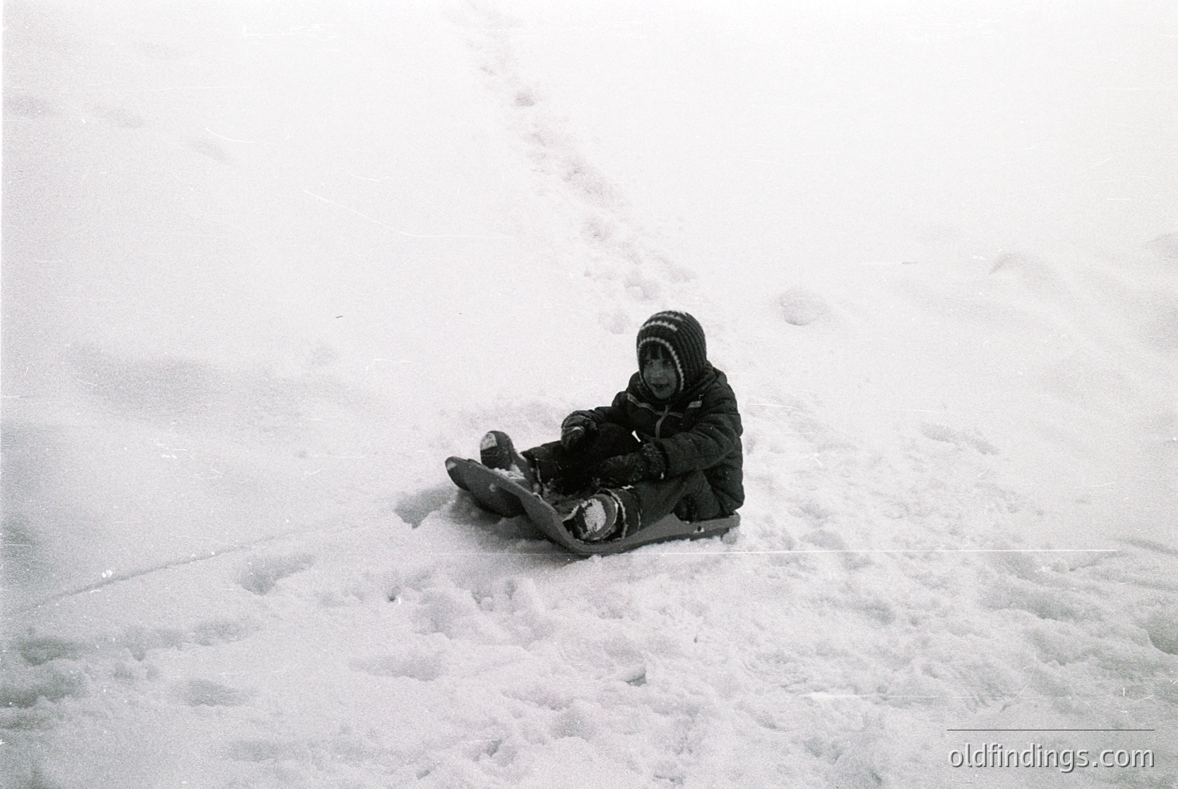 Black-and-white snapshot of a child sledding on a snow-covered slope, mid-20th century. The child wears a hooded jacket and sits on a small sled, facing forward. Snowy terrain suggests a recreational winter scene.