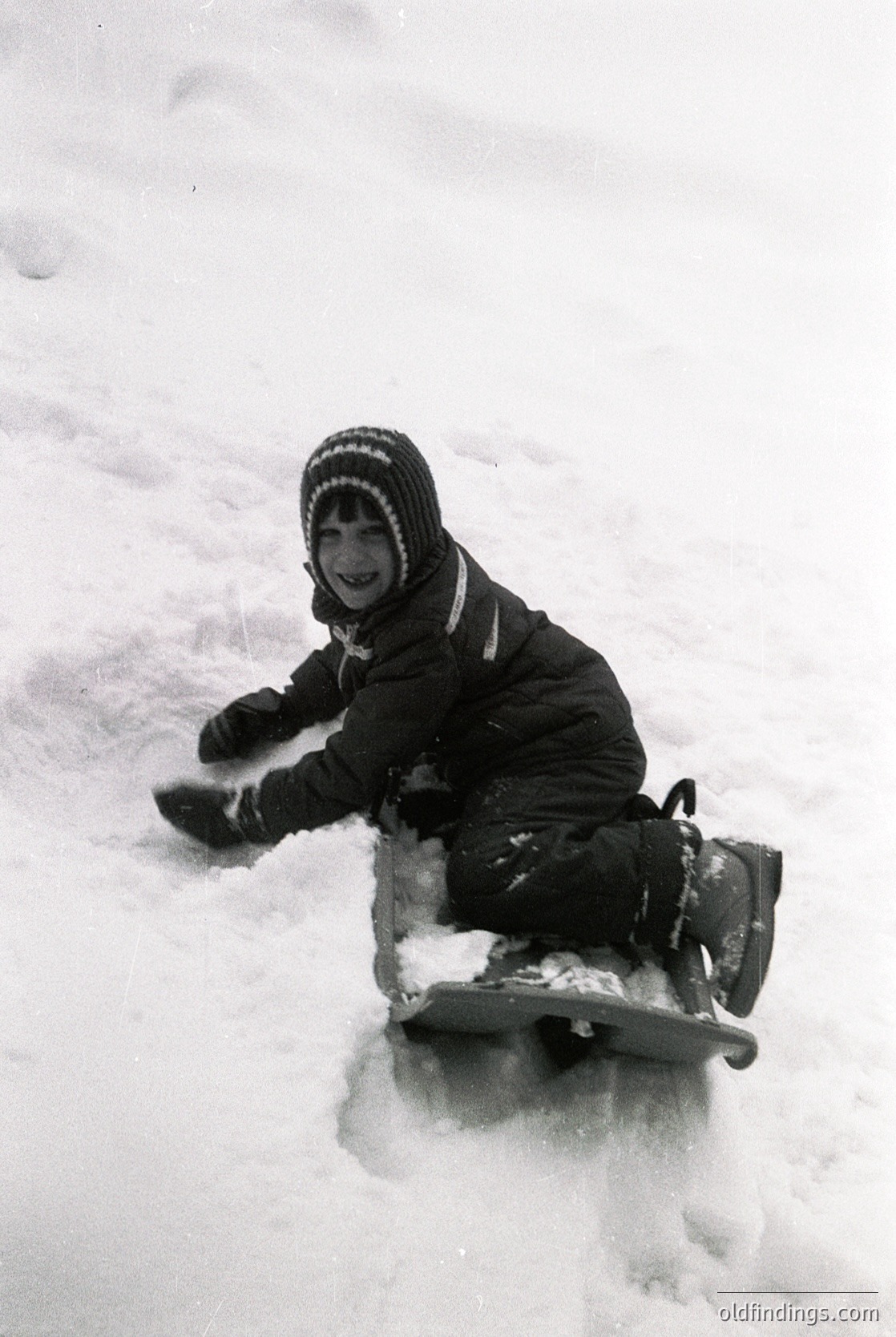 Child sledding on snow-covered ground, wearing a striped knit hat, dark winter coat, and mittens. Classic winter activity, likely mid-20th century.