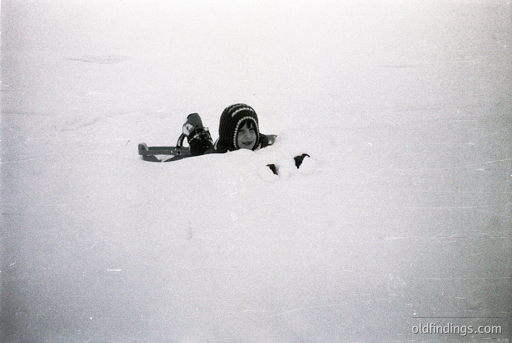 Black-and-white close-up of a vintage camera lens partially buried in snow, likely a **Leica-style rangefinder**. The composition highlights texture and contrast between the smooth metal and rough snow. Potential mid-20th century (1940s–1960s) photography reference.