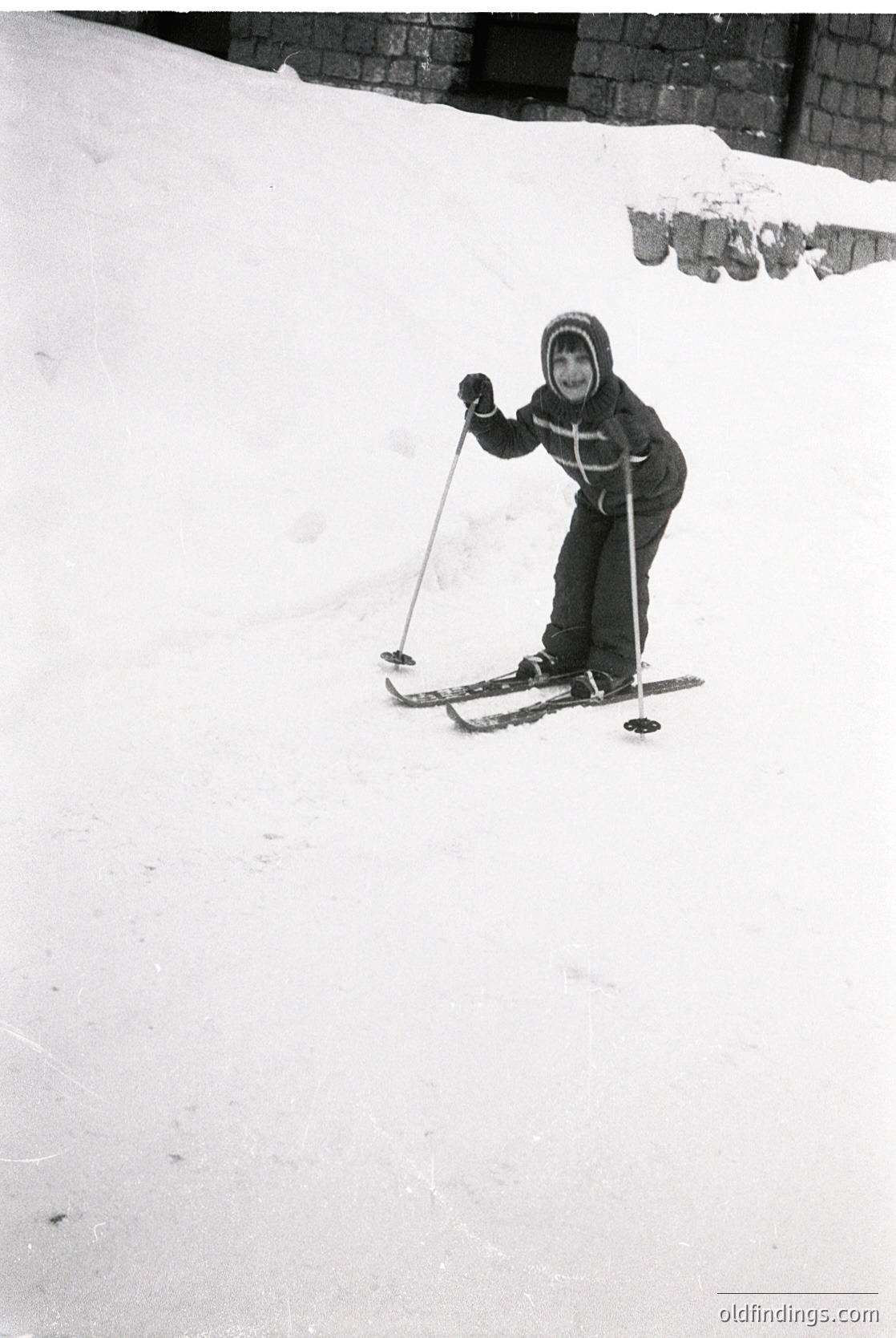 Child in vintage ski gear mid-turn on groomed slope, 1960s-70s style. Hooded jacket with cross patch, classic wooden skis, and metal poles. Snow-covered slope with brick wall in background.