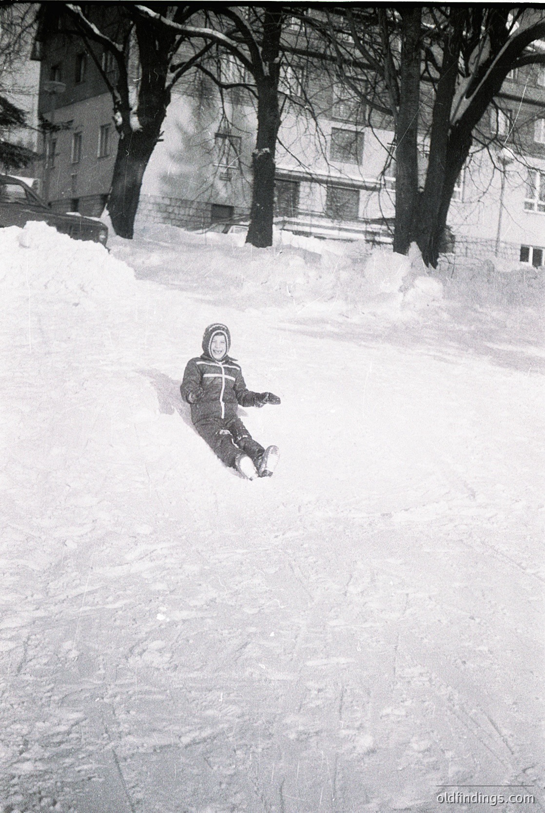Child sliding down snow-covered hill in urban park, mid-1970s. Snowy terrain contrasts with bare winter trees and multi-story residential buildings in background. Classic winter attire includes striped sweater and knit cap.