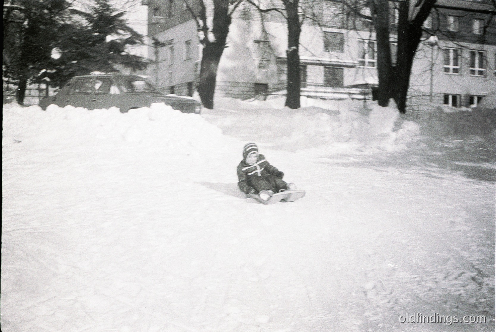 Child sledding on snow-covered urban slope, mid-20th century. Snow piles and parked vintage car in background suggest residential area. Black-and-white photo captures winter play and mid-century urban life.
