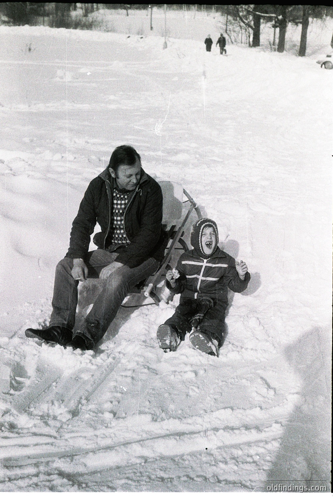 A man and child sledding in snow, mid-20th century. The man, seated, guides a child on a sled, both dressed in winter attire—checkered sweater, goggles, and boots. Snow-covered landscape with trees and distant figures in the background.